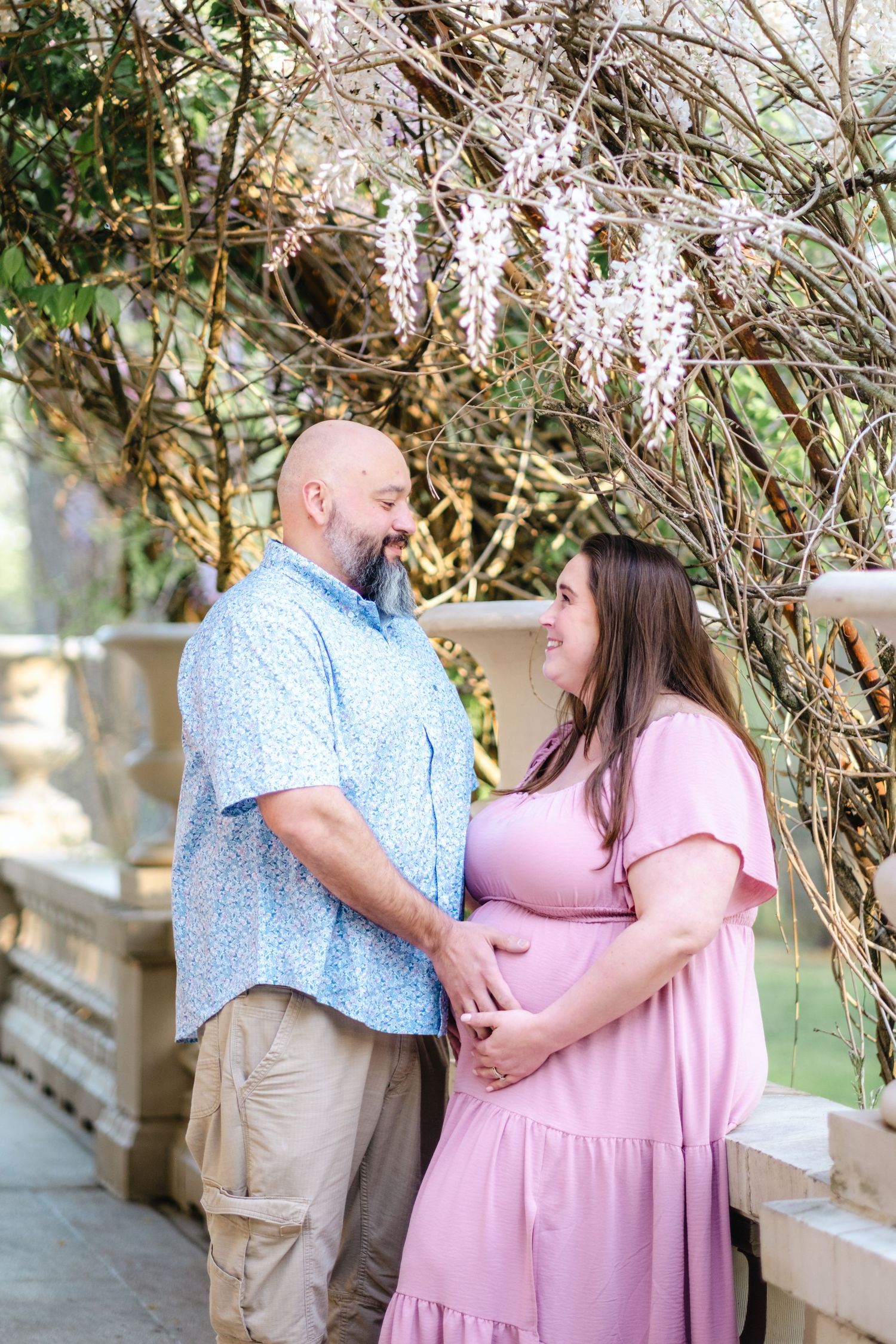 A couple shares a joyful moment outdoors, the woman in a pink maternity dress cradling her baby bump under blooming wisteria.