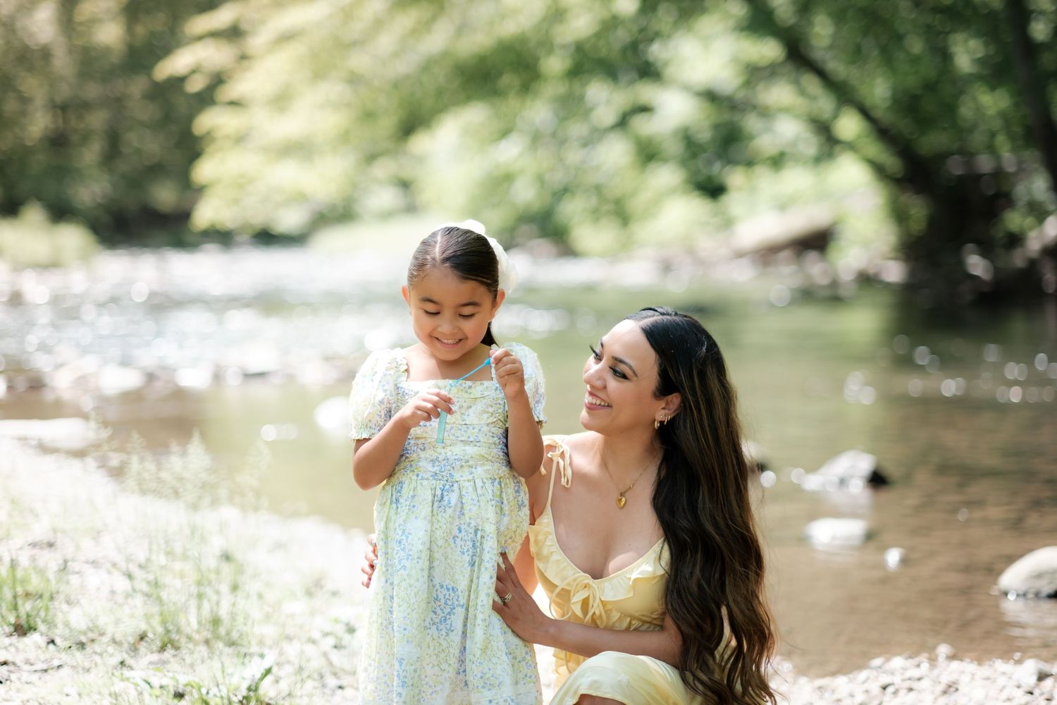 A mother kneels beside her young daughter in a floral dress near a sunlit creek surrounded by lush green trees.