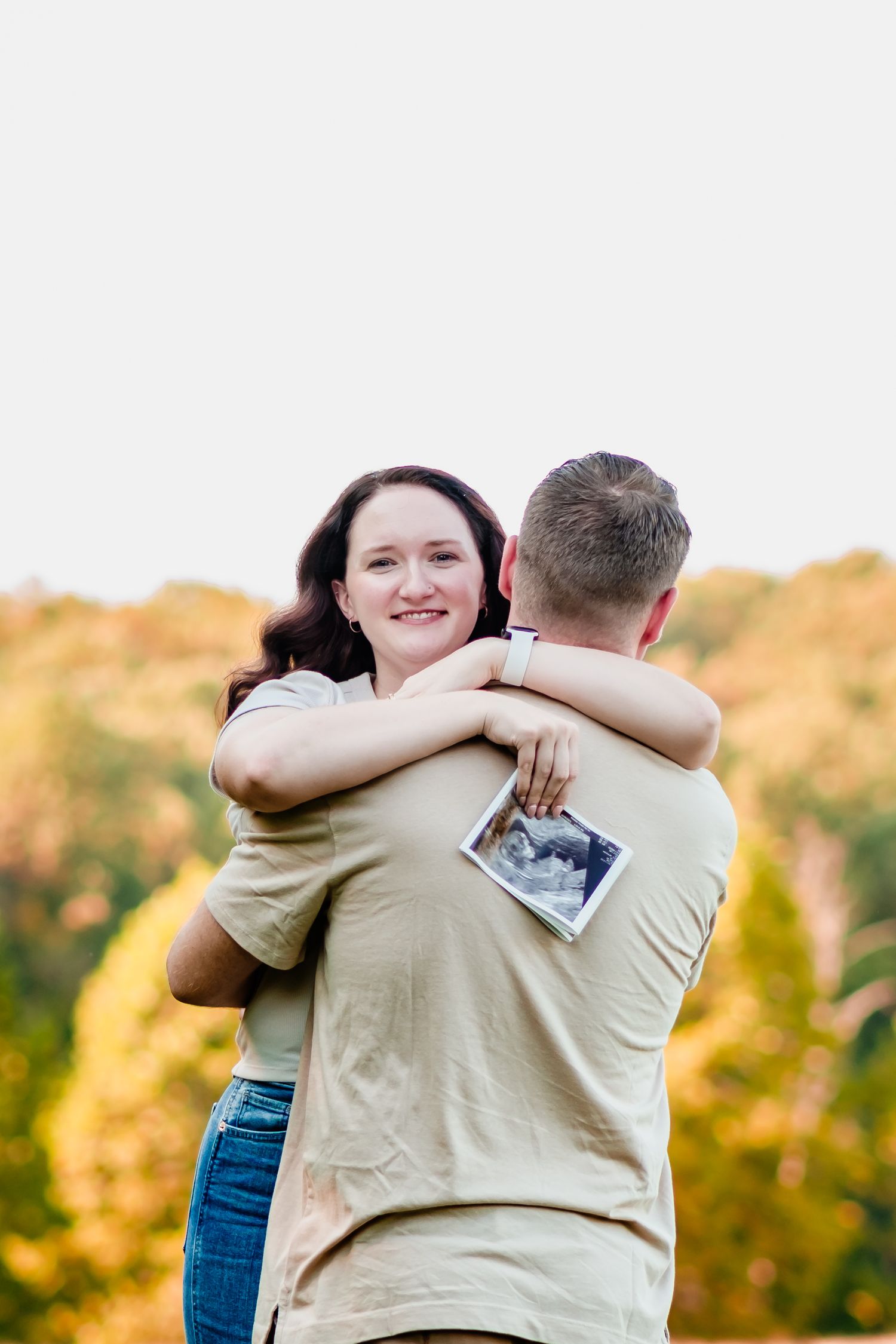 Couple embracing outdoors in fall, woman smiling and holding ultrasound photo, announcing pregnancy.