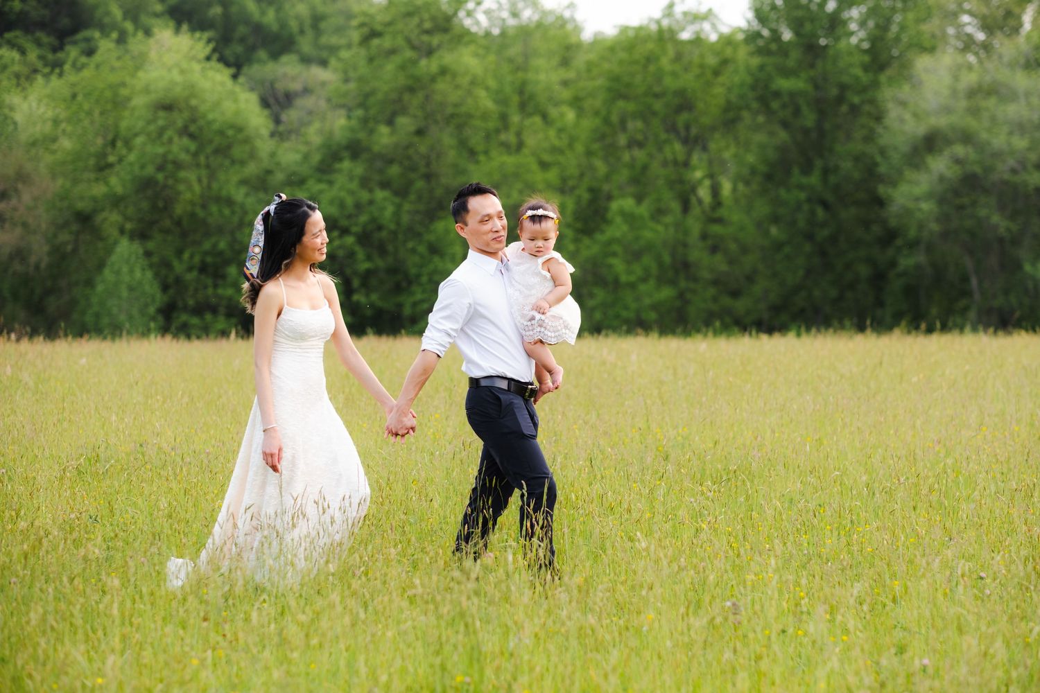 Bride and groom walking hand-in-hand in a green meadow, groom holding a baby, with lush trees in the background.