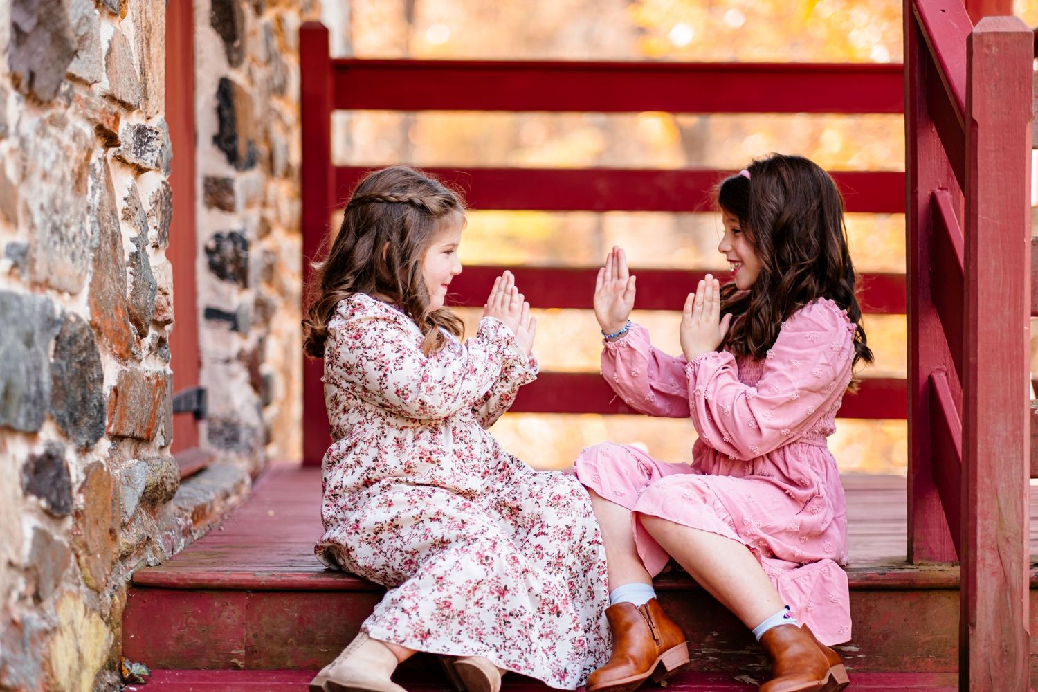 Two young girls in floral dresses playing patty-cake on red wooden steps near a stone wall.