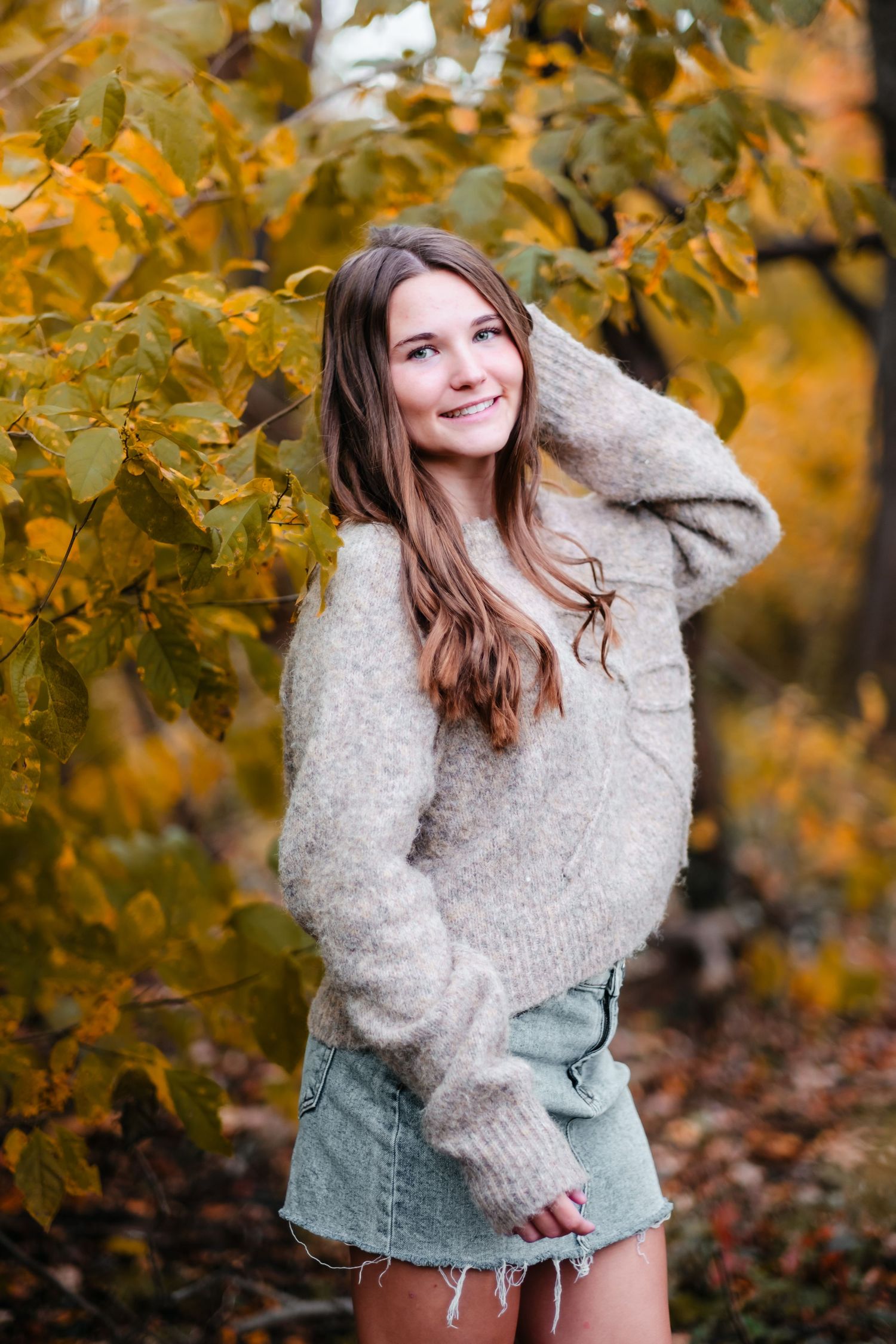 Young woman in cozy knit sweater and denim skirt smiling among vibrant autumn foliage in a fall outdoor setting.