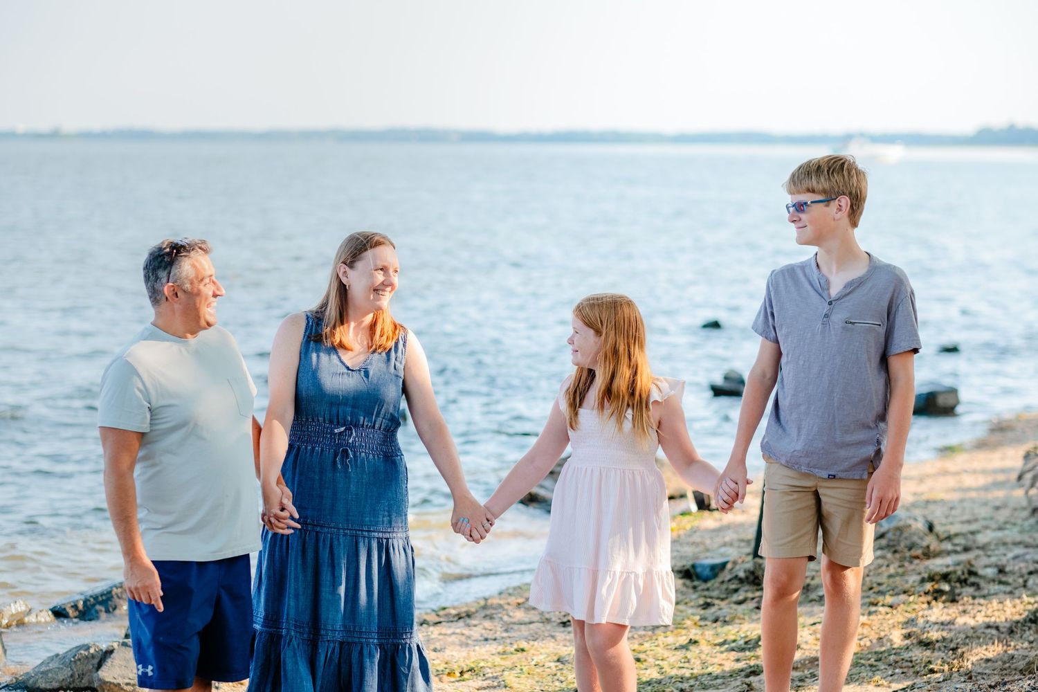 Family of four holding hands on a rocky beach shore, smiling on a sunny day near calm blue water.