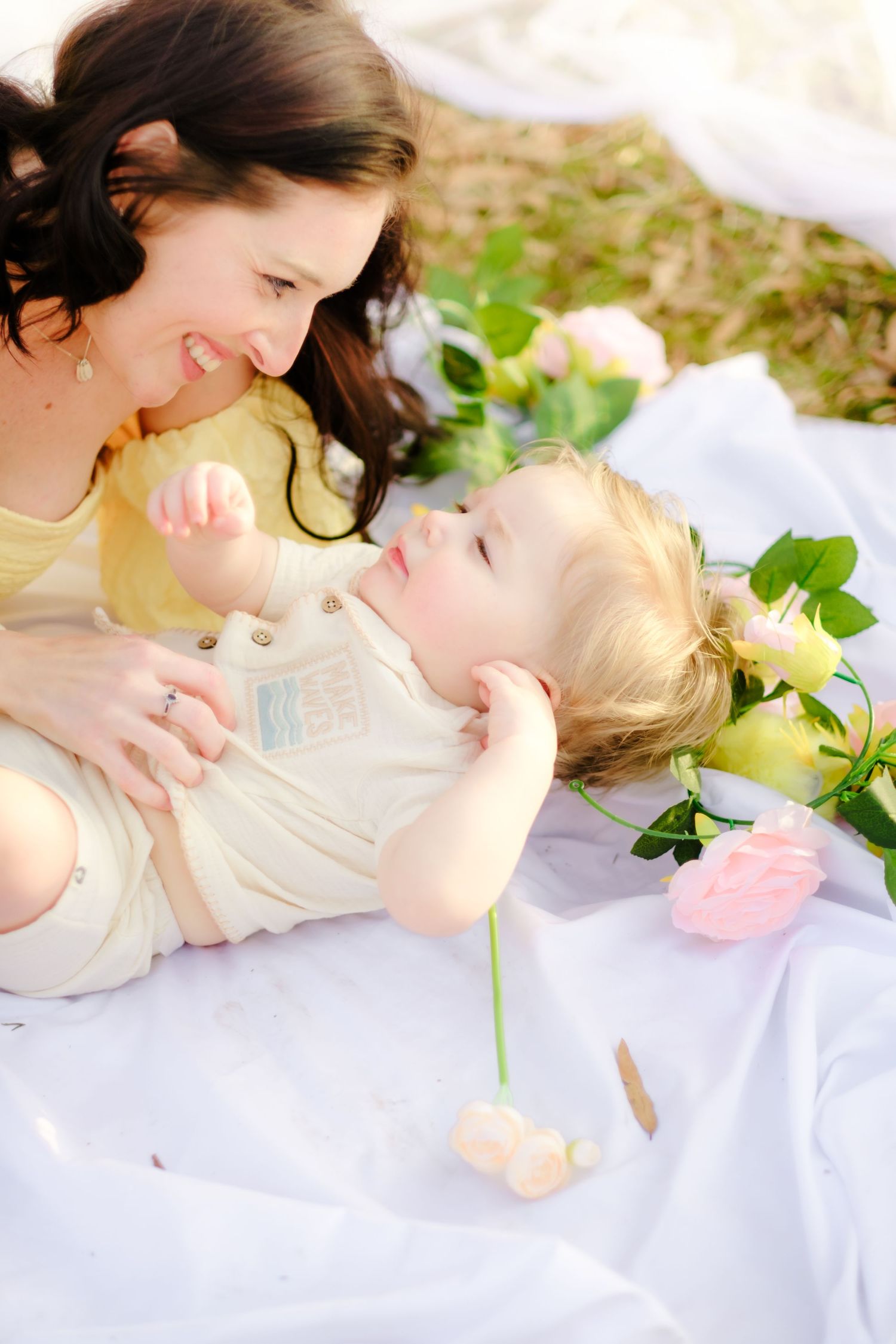 Joyful mother in yellow dress plays with smiling toddler lying on white blanket surrounded by pink roses.
