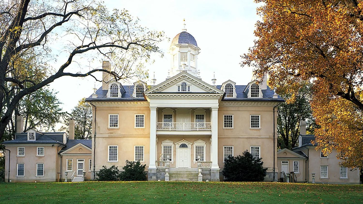 Historic yellow Georgian mansion with white columns, cupola, and balcony surrounded by autumn trees on manicured lawn.