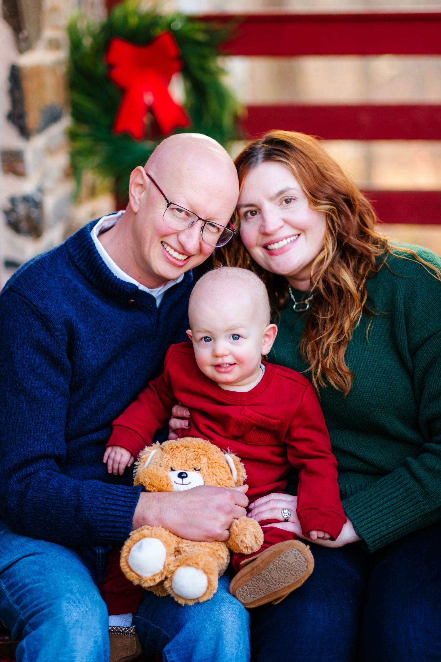 Family Christmas portrait with parents and baby holding teddy bear, festive holiday decor in background.