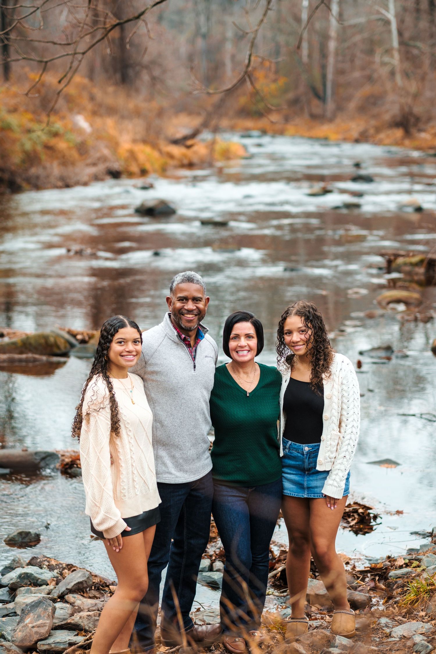 Family of four smiling together by a rocky stream surrounded by autumn foliage in a serene outdoor setting.