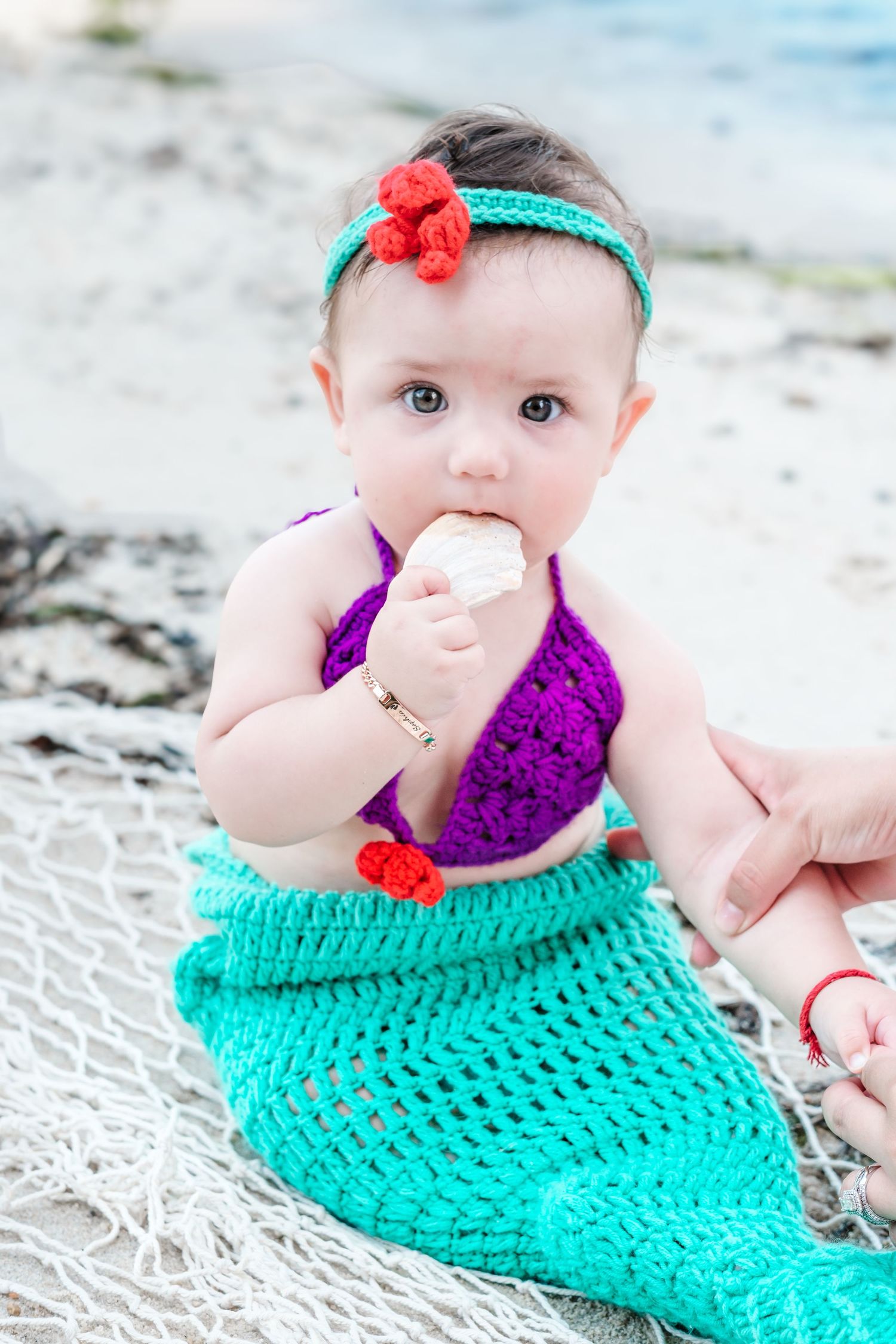 Baby girl in crochet mermaid costume holding a seashell on a beach.