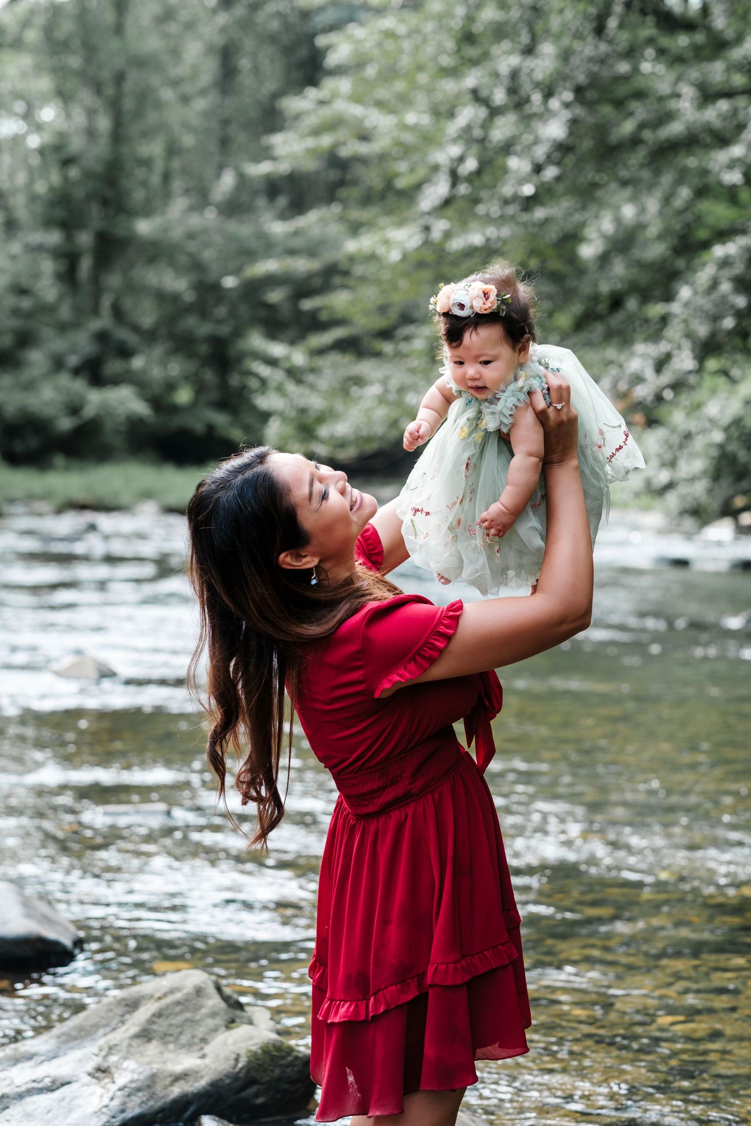 A woman in a red dress holds a baby girl in a floral outfit above a rocky stream surrounded by green trees.