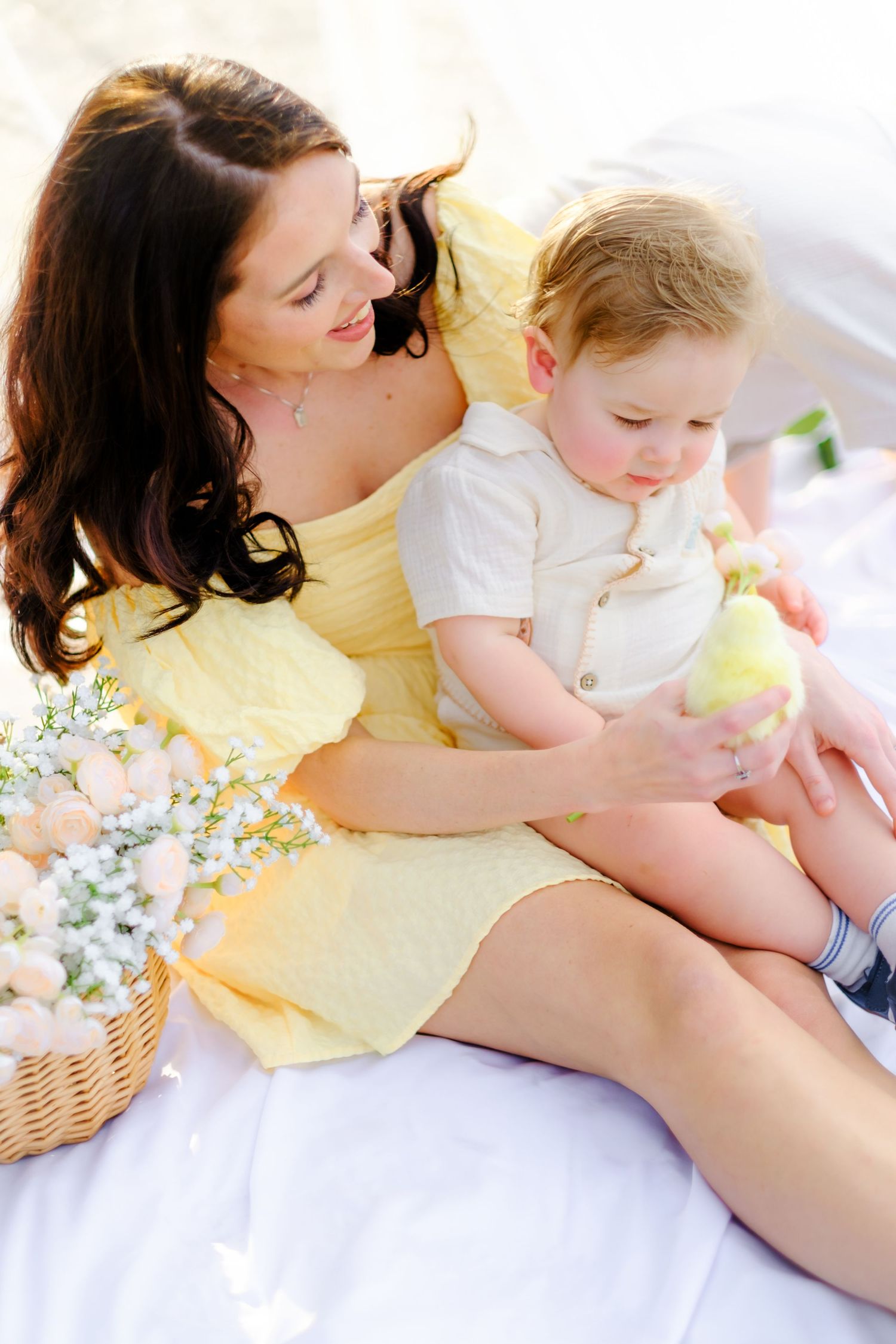 Mother in yellow dress holds toddler son on white blanket during outdoor spring picnic with flower basket.