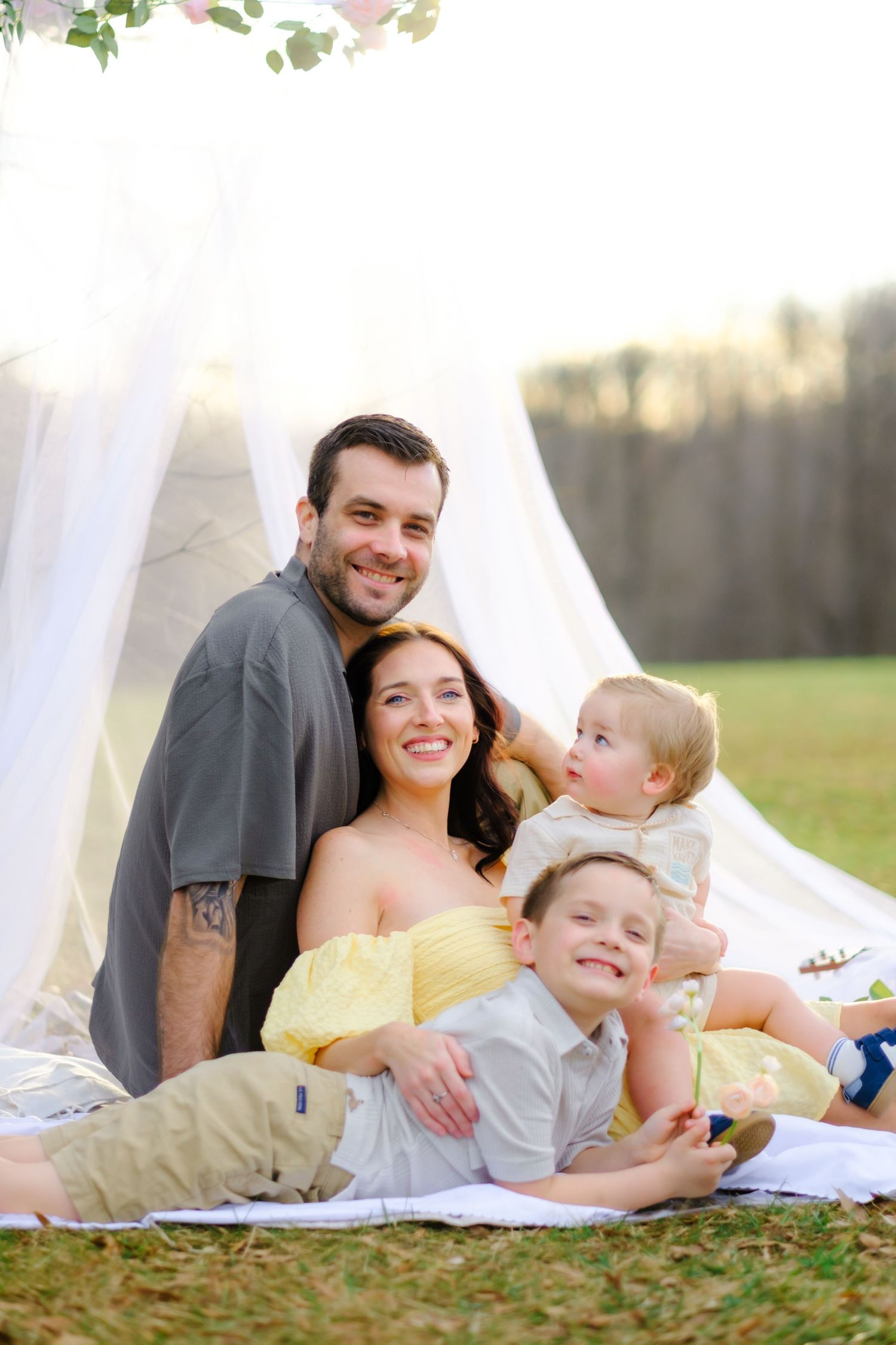 Happy family of four posing outdoors on a blanket near a white canopy with floral decor on a sunny day.