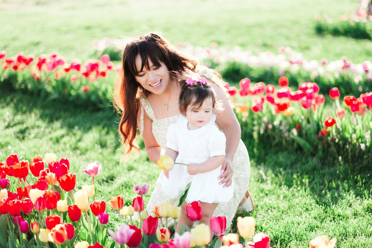 Mother and baby girl in white dresses smiling among colorful red and pink tulips in a sunny garden.