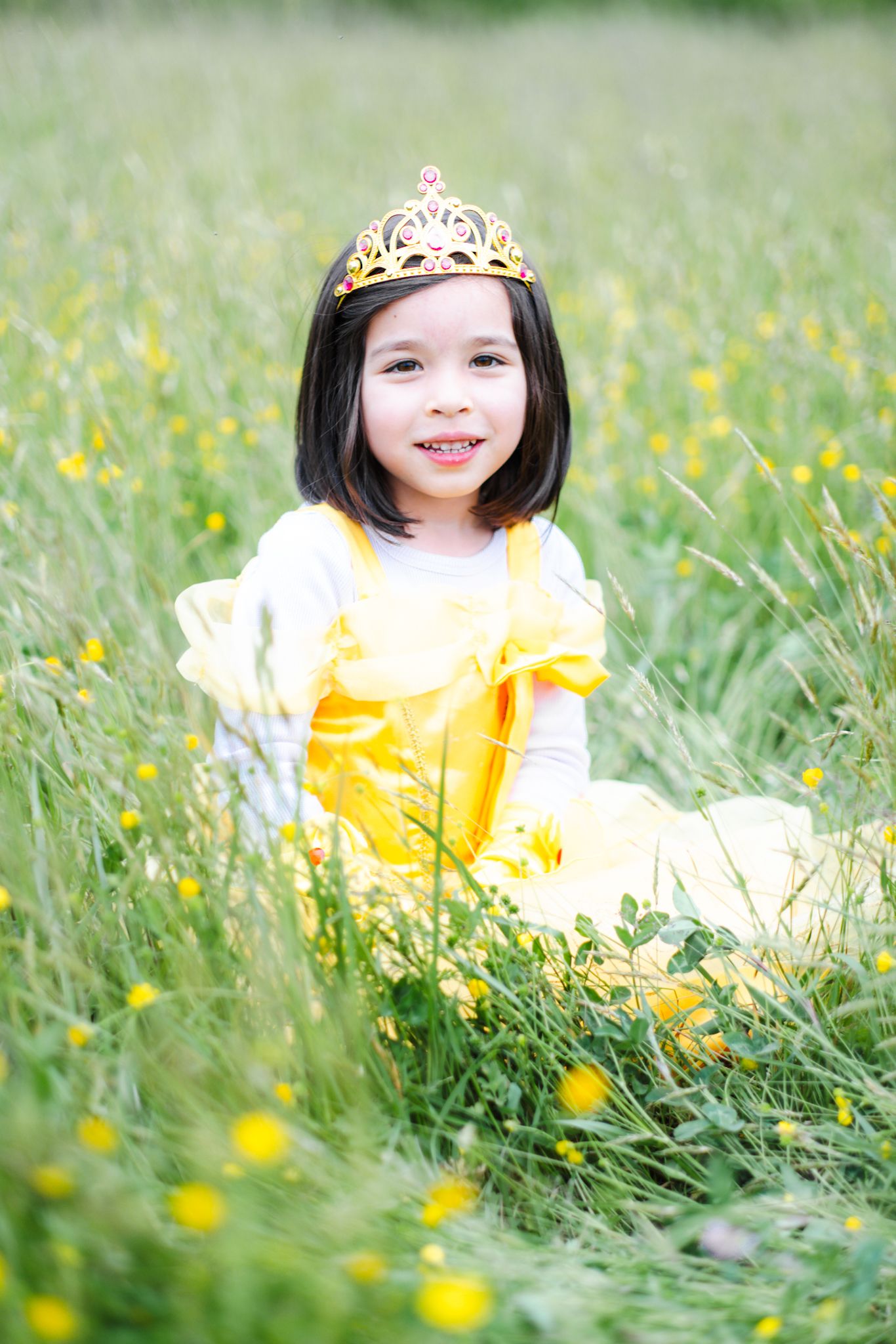 A smiling little girl in a yellow princess dress and gold tiara sits in a green meadow with yellow wildflowers.