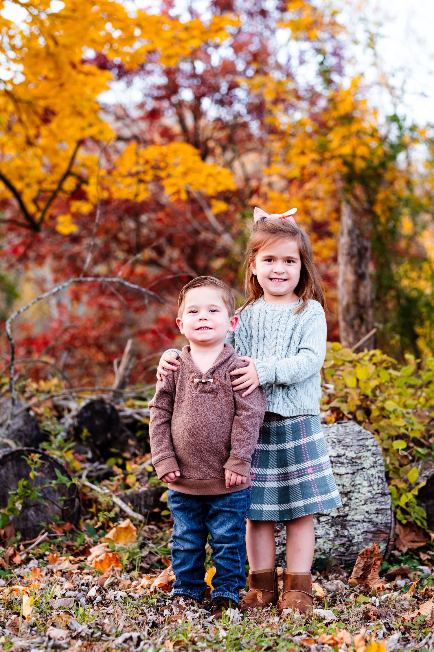 Two young siblings pose together outdoors among vibrant autumn foliage, girl in plaid skirt hugging younger boy in brown sweater.