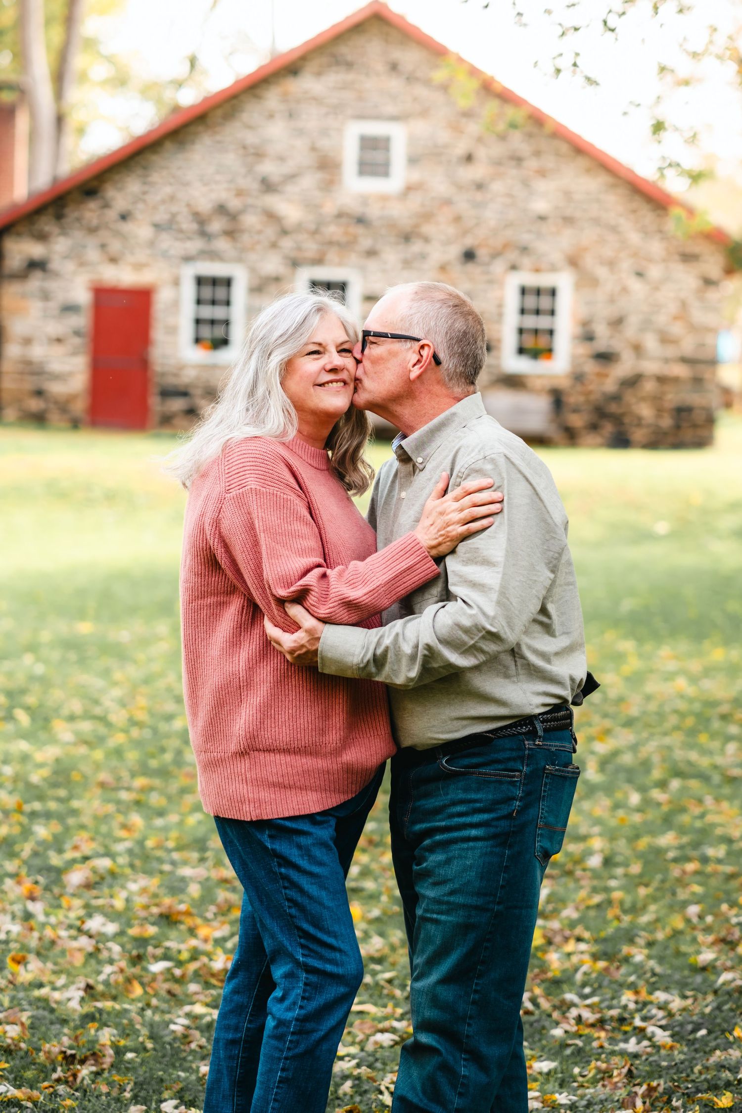 An older couple sharing a romantic kiss outdoors in autumn, standing in front of a rustic stone building.