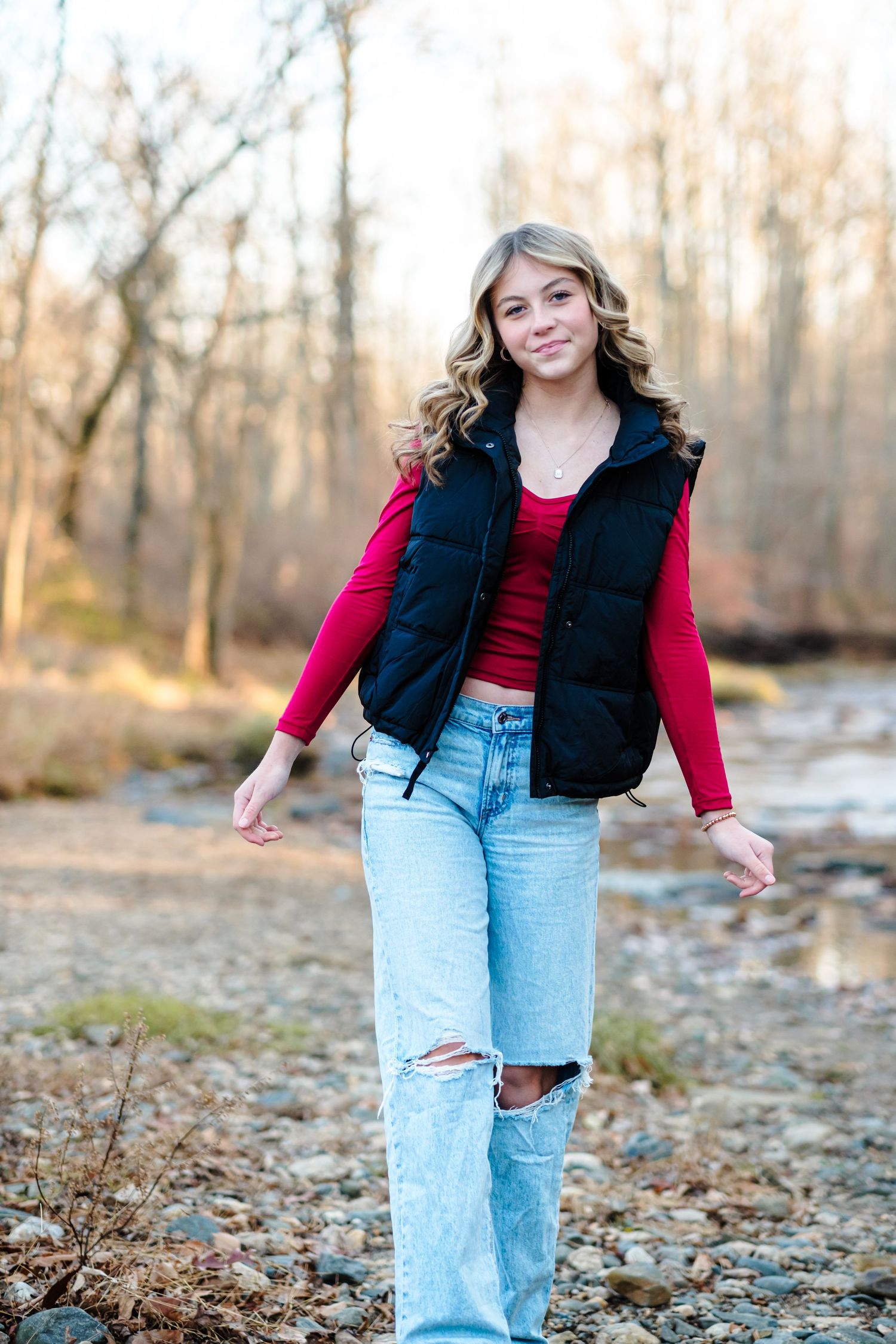 Smiling teenage girl walking by a rocky creek wearing a black vest, red long-sleeve top, and ripped jeans.