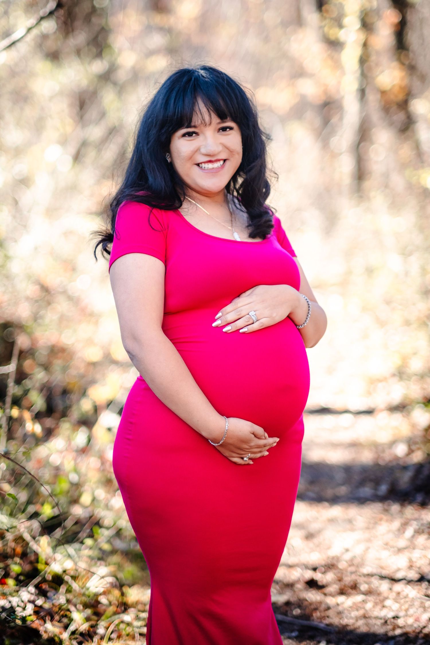 Smiling pregnant woman in pink dress cradling baby bump outdoors in a sunlit forest setting.