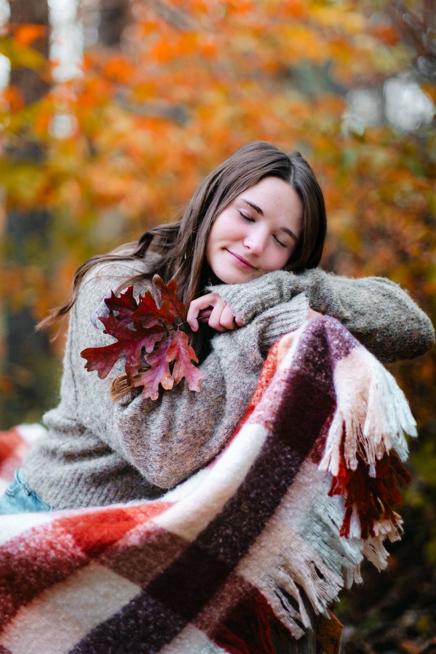 Young woman with closed eyes hugging a plaid blanket and autumn leaves in a cozy fall forest setting.