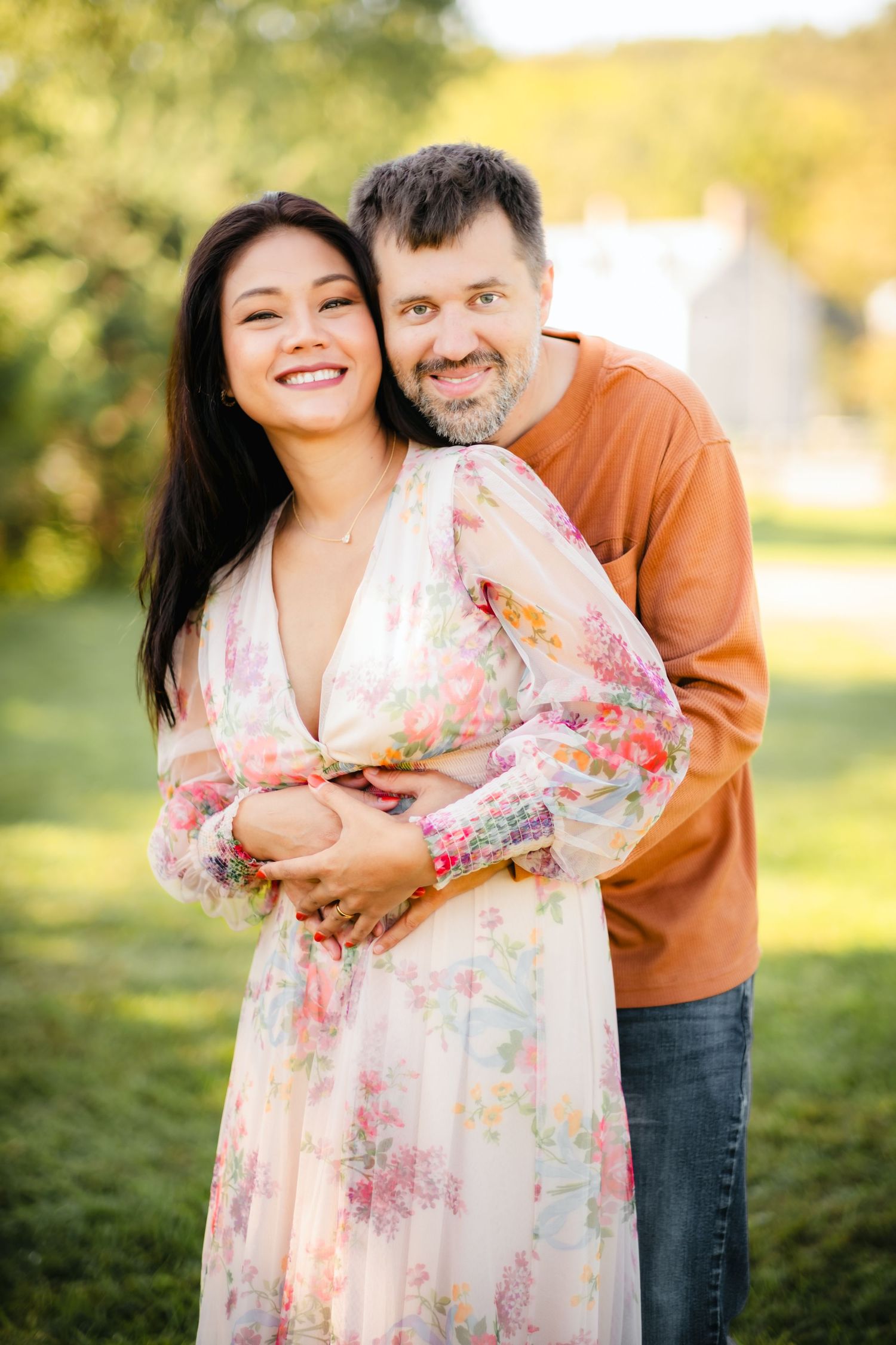 Happy couple embracing outdoors, woman in floral dress and man in orange shirt, smiling in a lush green park setting.