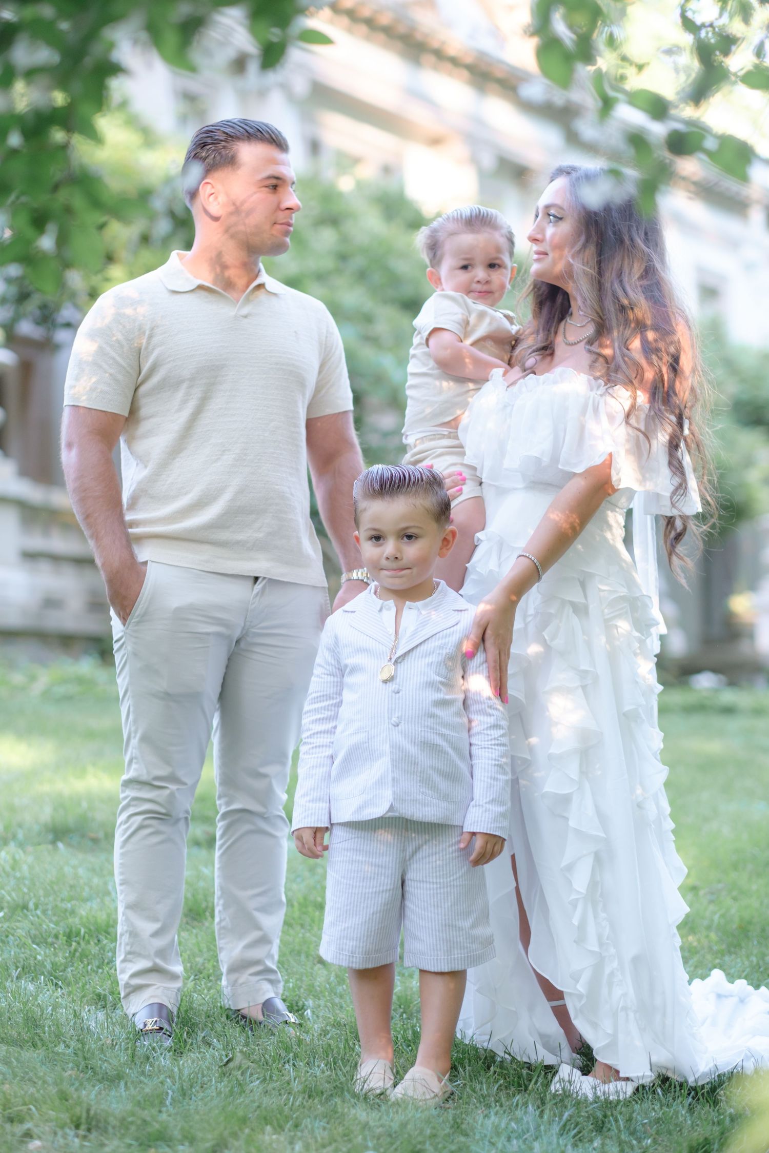 A happy family of four dressed in white and beige outfits poses outdoors on green grass near trees.