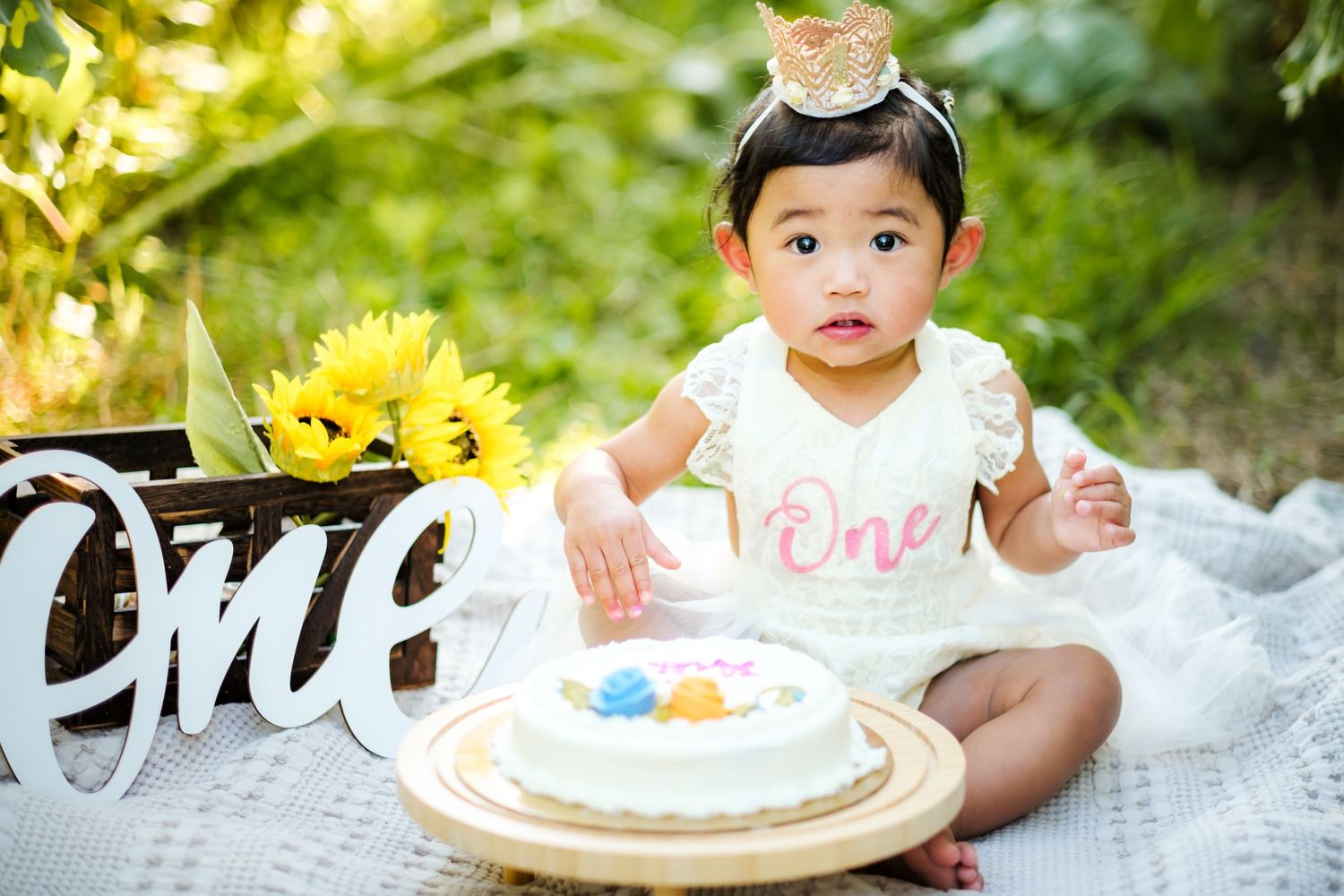 Baby girl in white dress with crown celebrates first birthday with cake outdoors among sunflowers.