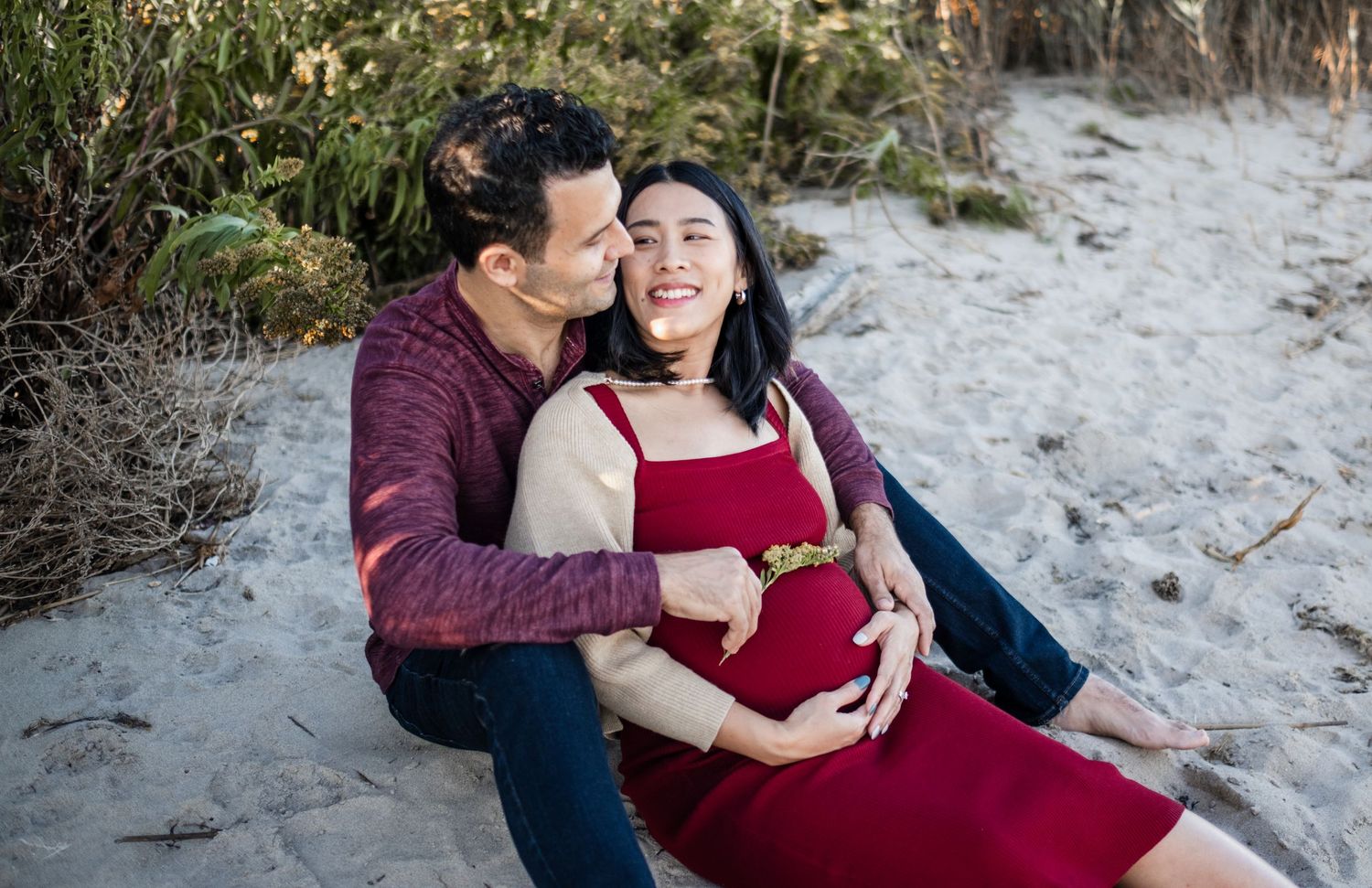 Couple sharing a tender maternity moment on a sandy beach, woman in red dress cradling baby bump.
