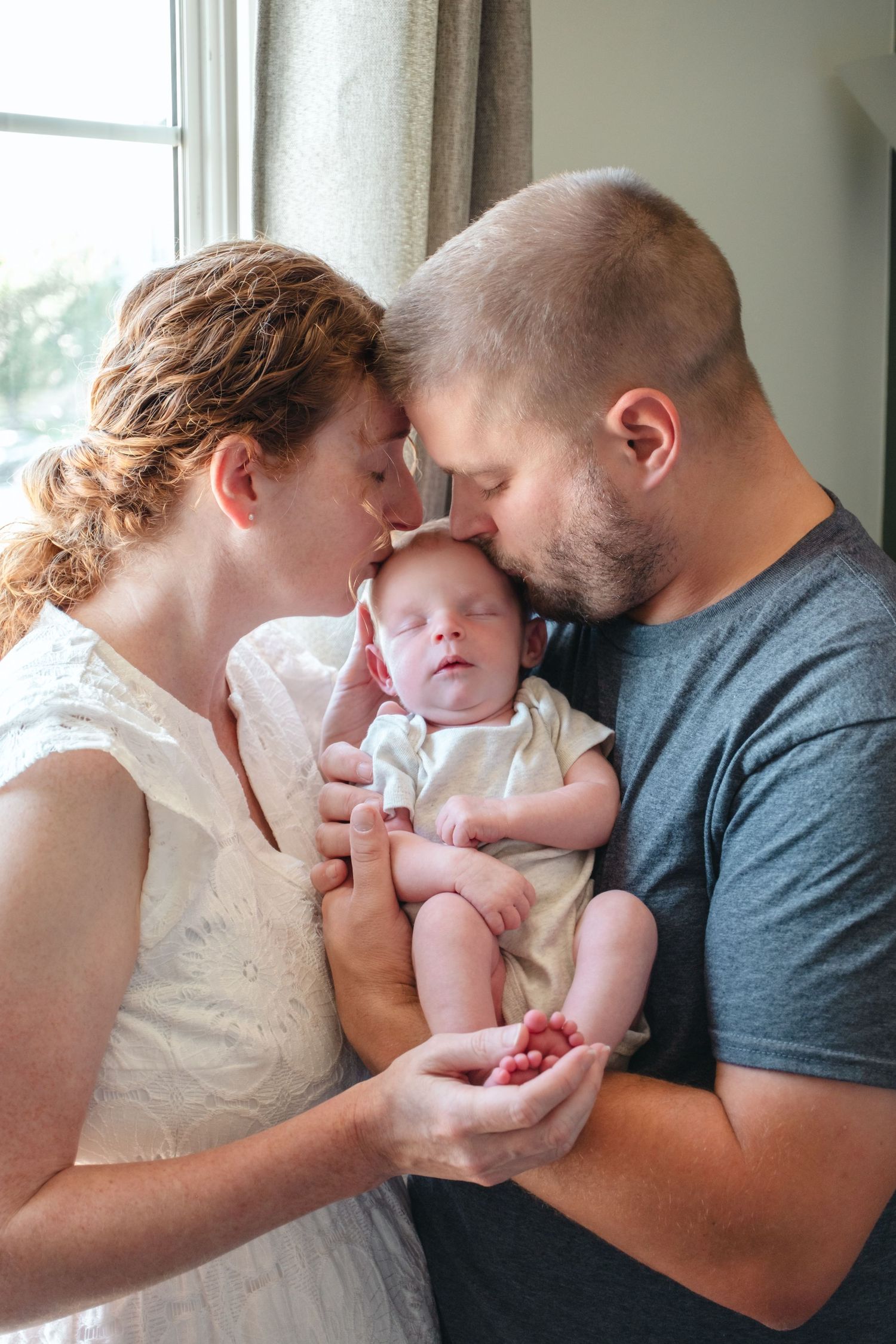 New parents tenderly touch foreheads while holding their sleeping newborn baby near a bright window.