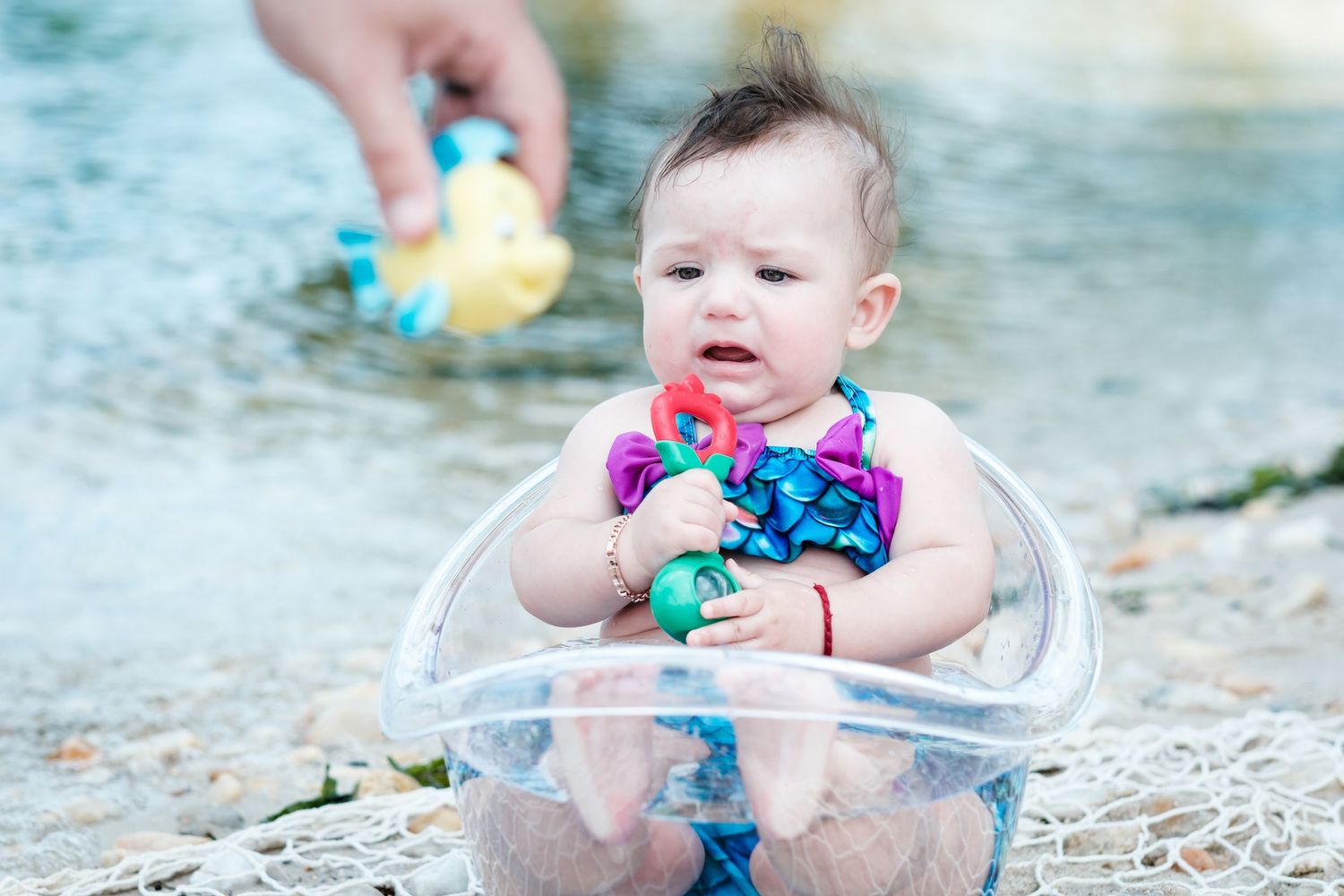 Cute baby sitting in a clear bowl on a beach, holding colorful toys while an adult offers a rubber duck.