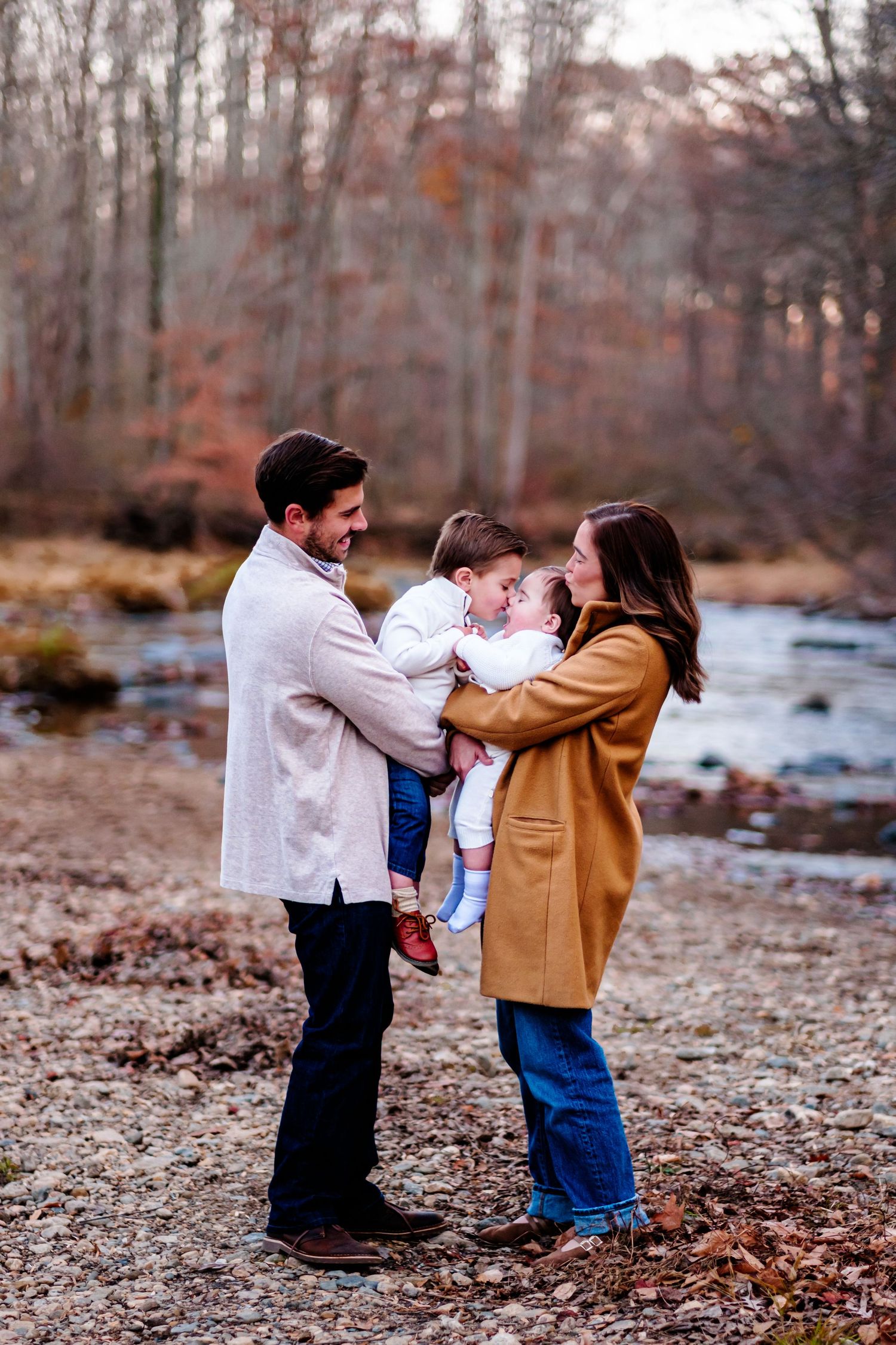 A happy family of three poses by a creek in autumn woods, parents holding their toddler and smiling warmly.