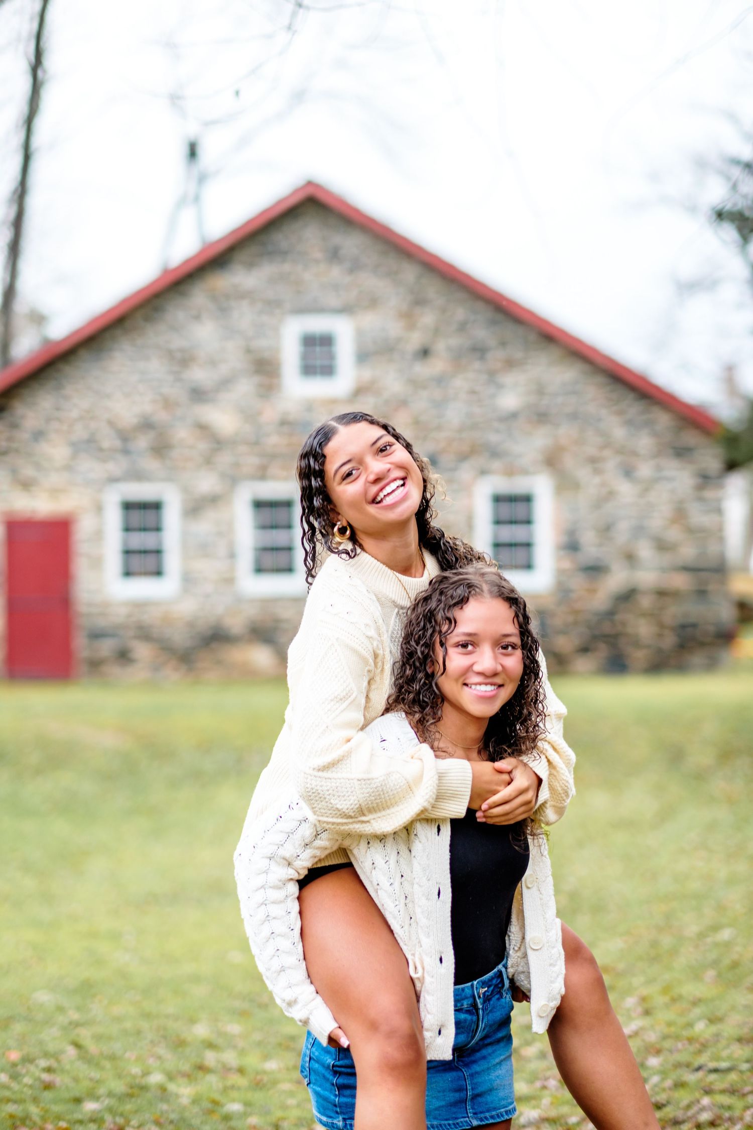 Two smiling young women pose outdoors, one giving the other a piggyback ride near a rustic stone building.