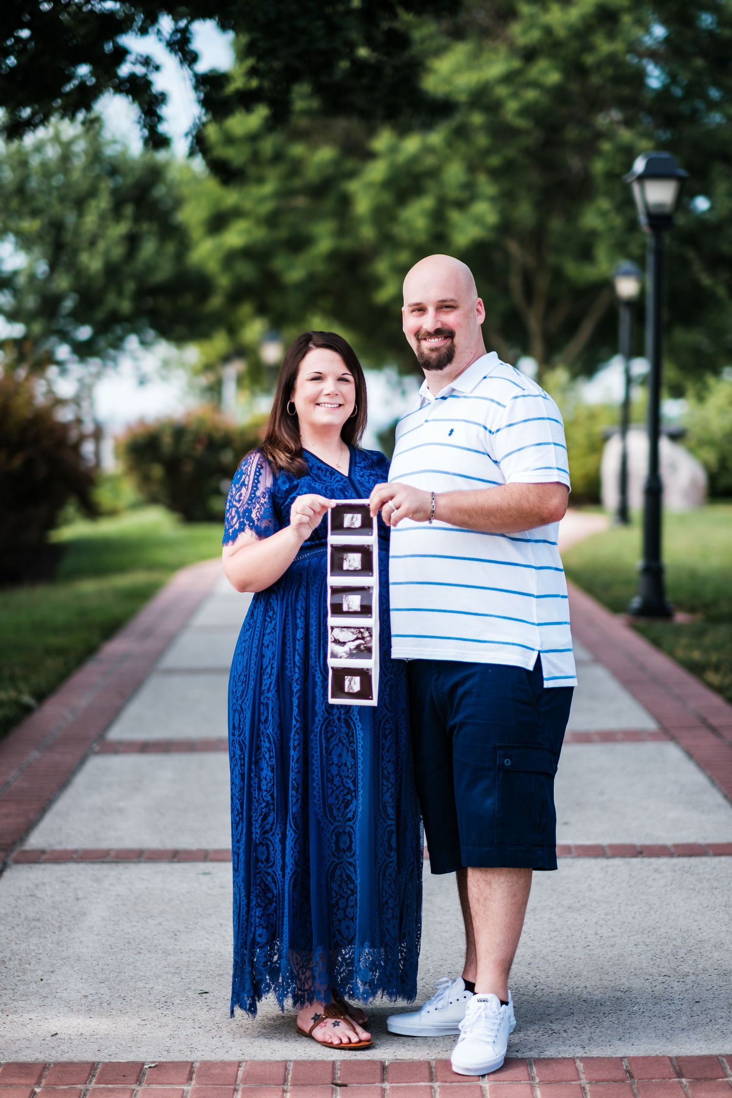 Happy couple holding ultrasound photos outdoors on a brick pathway, announcing their pregnancy in a lush green park setting.