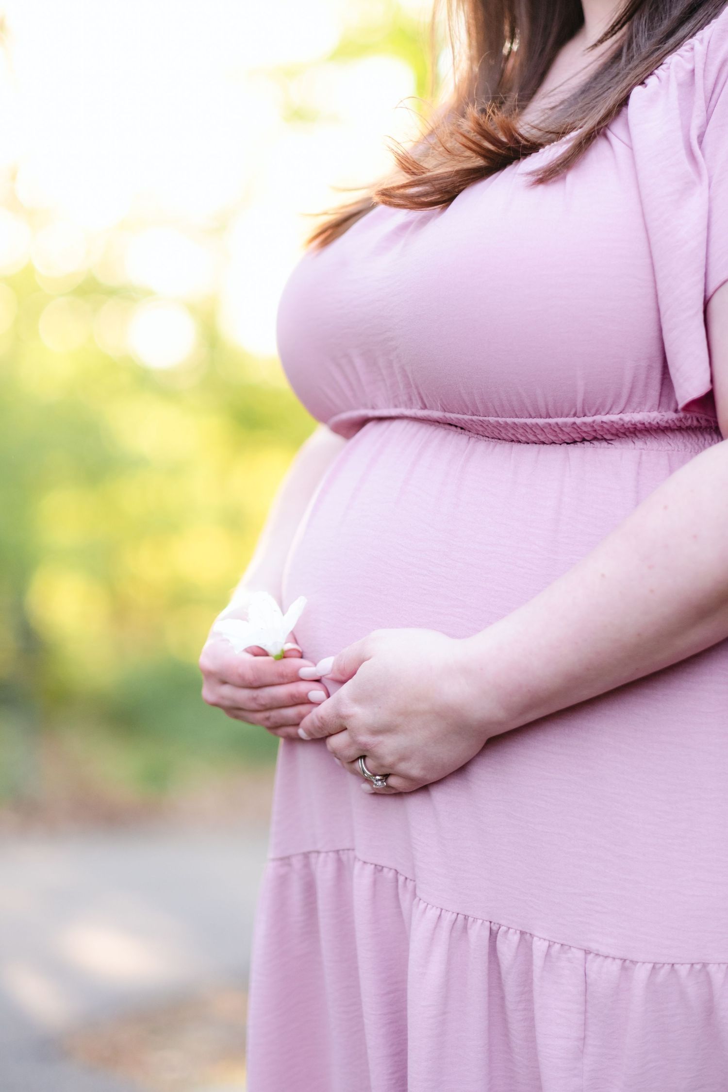 Pregnant woman in pink dress cradling baby shoes against her baby bump outdoors.