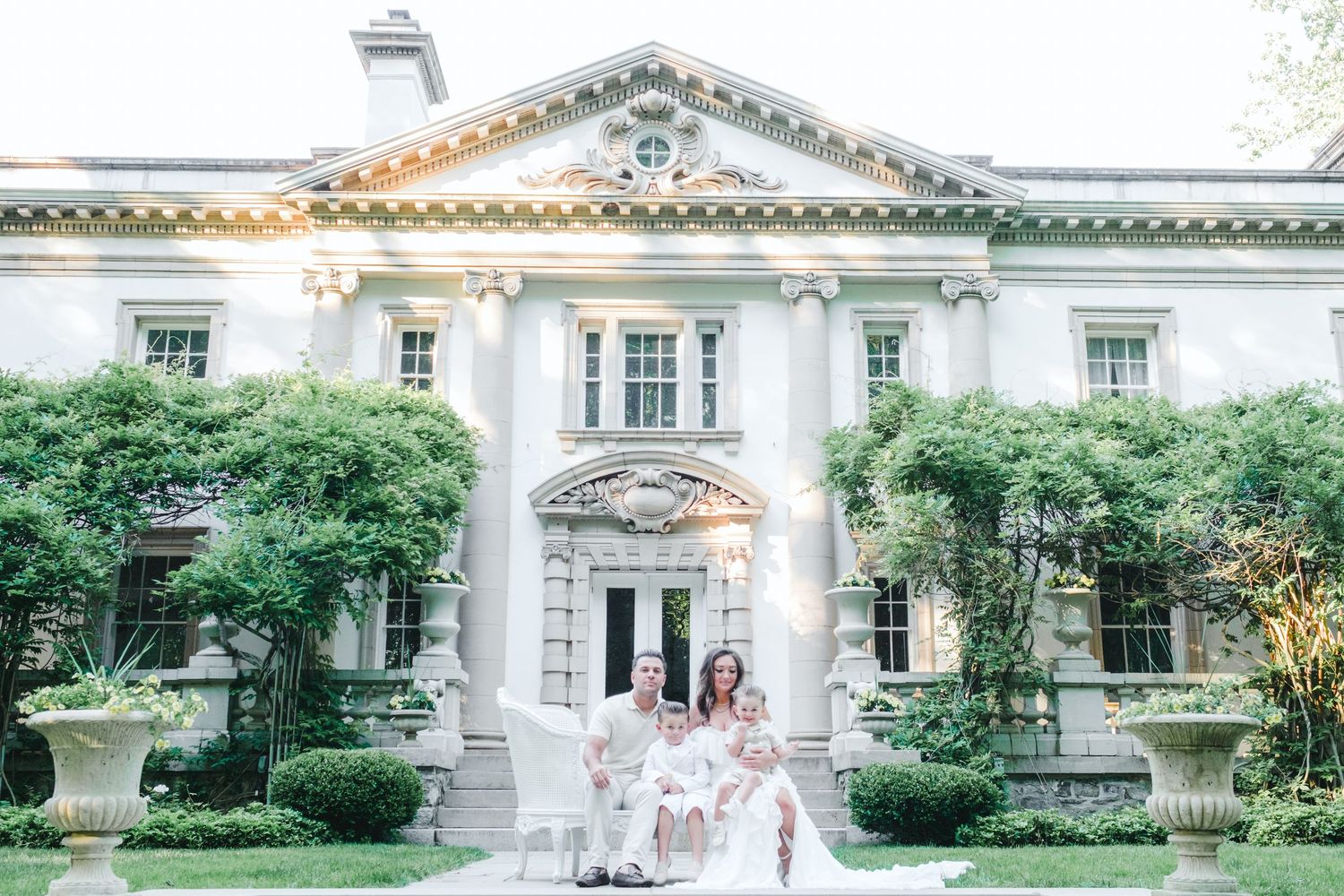 Bridal party posing on steps of elegant neoclassical mansion with lush greenery and white columns.
