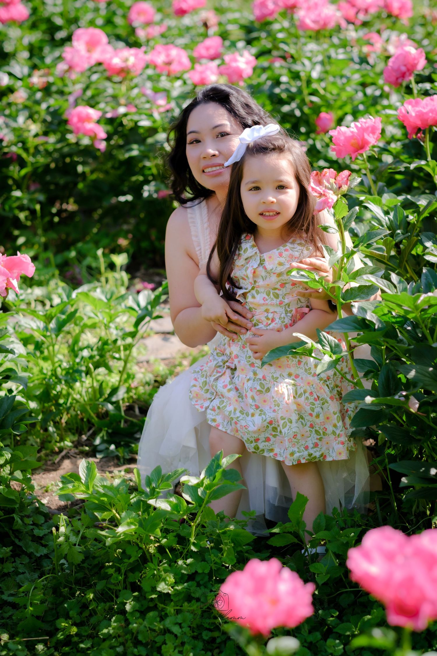 Mother and daughter posing together in a blooming pink rose garden, both wearing floral dresses on a sunny day.