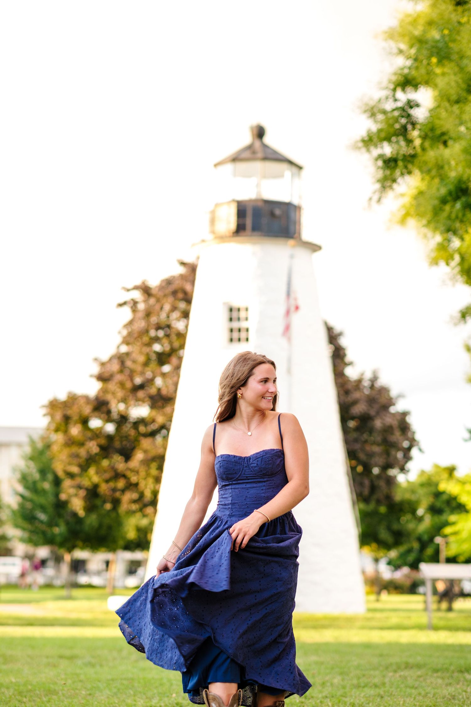 Woman in blue dress twirling near a white lighthouse on a sunny day.