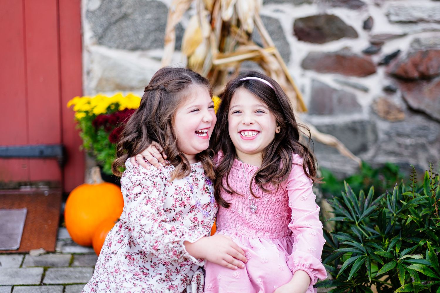 Two young girls laughing and hugging outdoors in fall setting with pumpkin and stone wall backdrop.