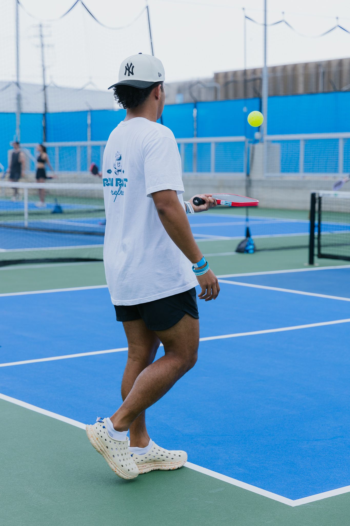 A tennis player wearing white shoes and shorts prepares to return a shot on a bright blue outdoor tennis court.