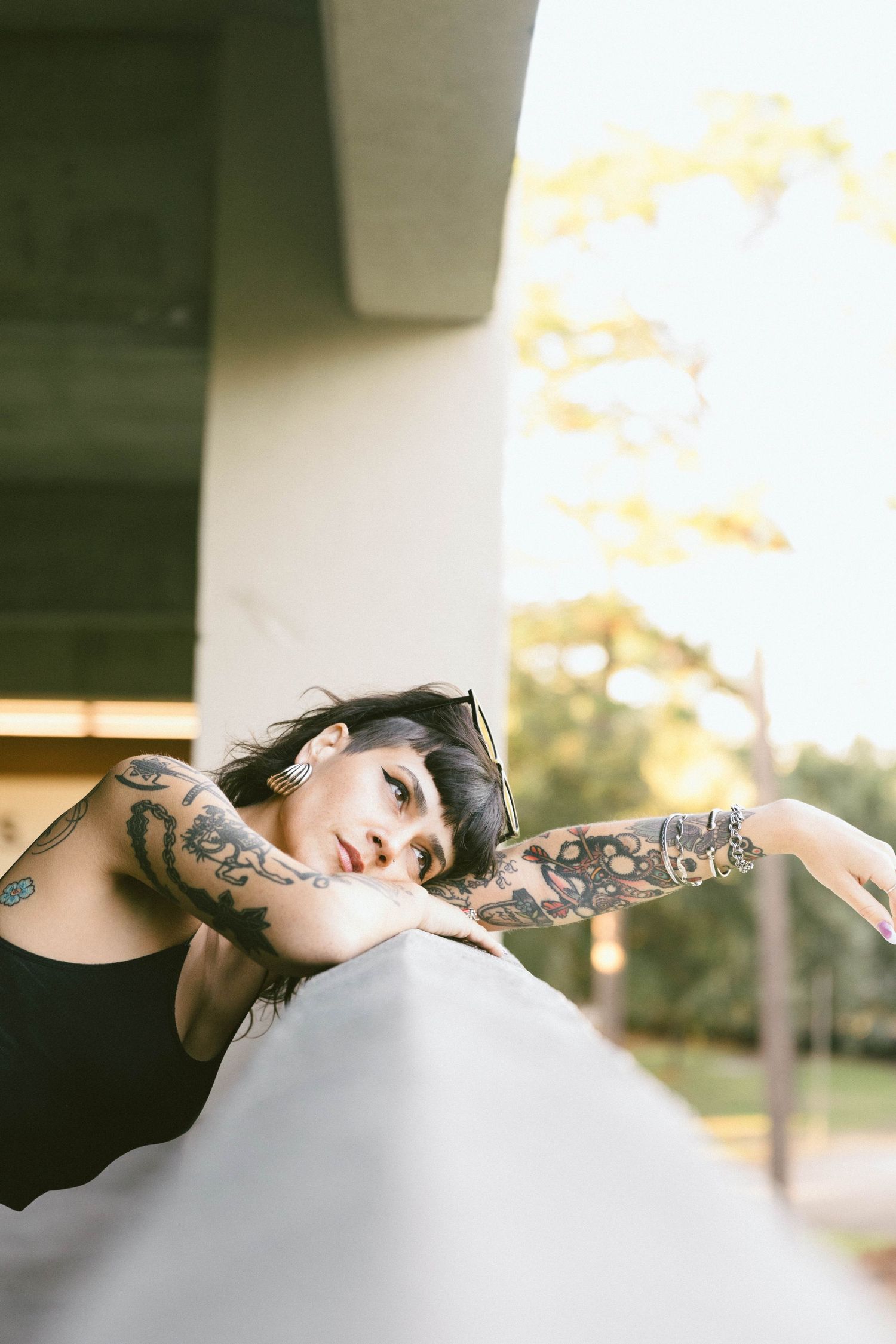 Person with tattoos leaning on a railing at sunset, wearing a black tank top in an outdoor setting.