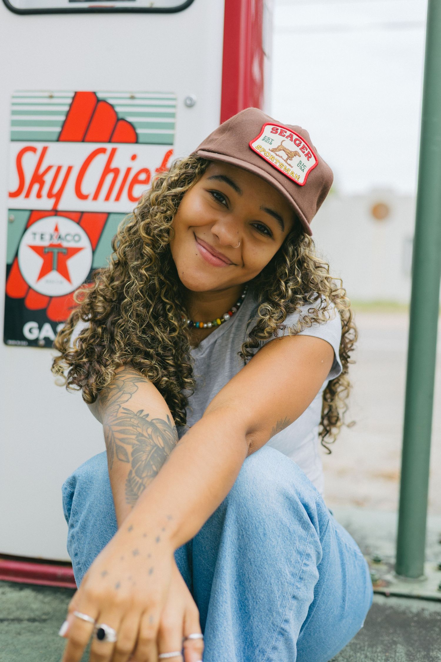 Person in red baseball cap and blue jeans poses casually in front of vintage gas station signs and decor.