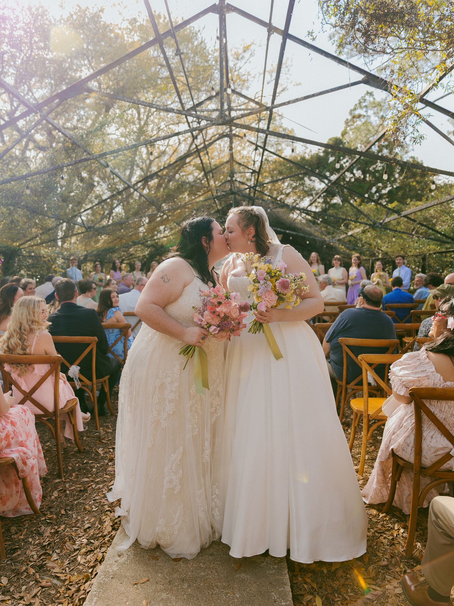 Two brides in white gowns share a first kiss at an outdoor wedding ceremony under a glass greenhouse structure.