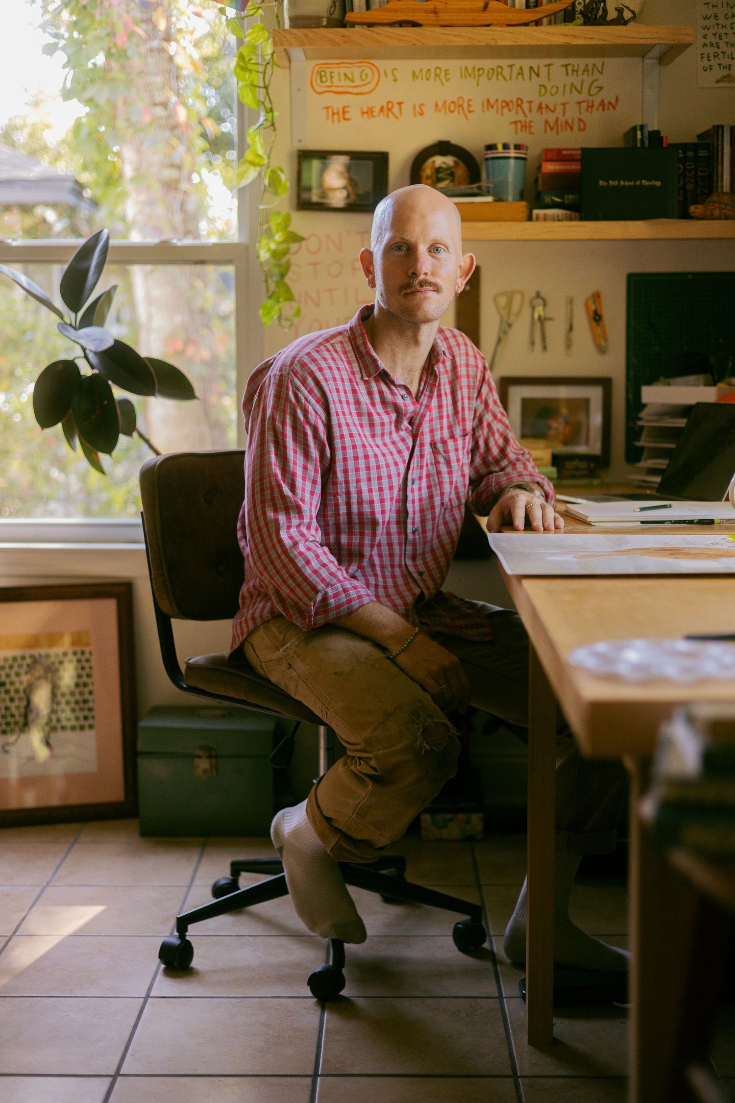 Artist sits at desk in sunlit home studio with plants, wearing plaid shirt and working at creative workspace with sketches on wall.