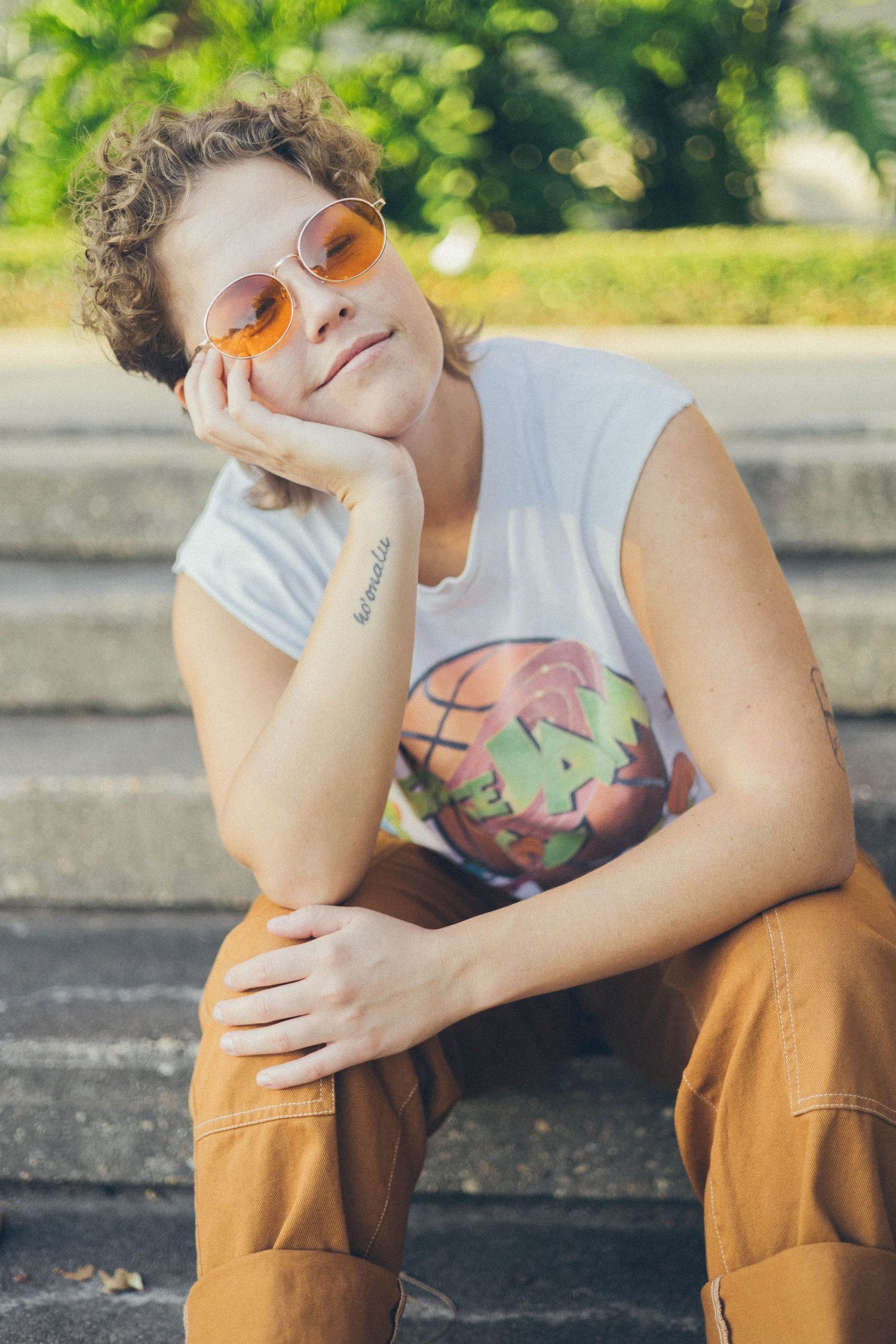 Person in orange sunglasses and brown pants sits thoughtfully on concrete ledge against green foliage background.