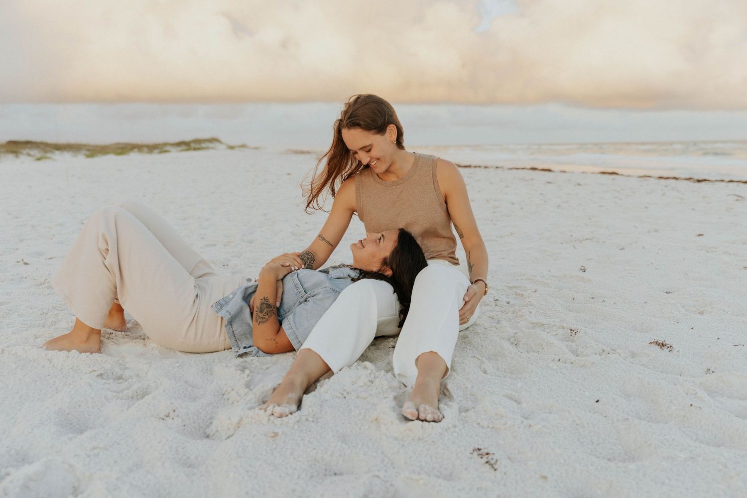 A couple sits close together on a white sandy beach at sunset wearing casual beige and white clothing.