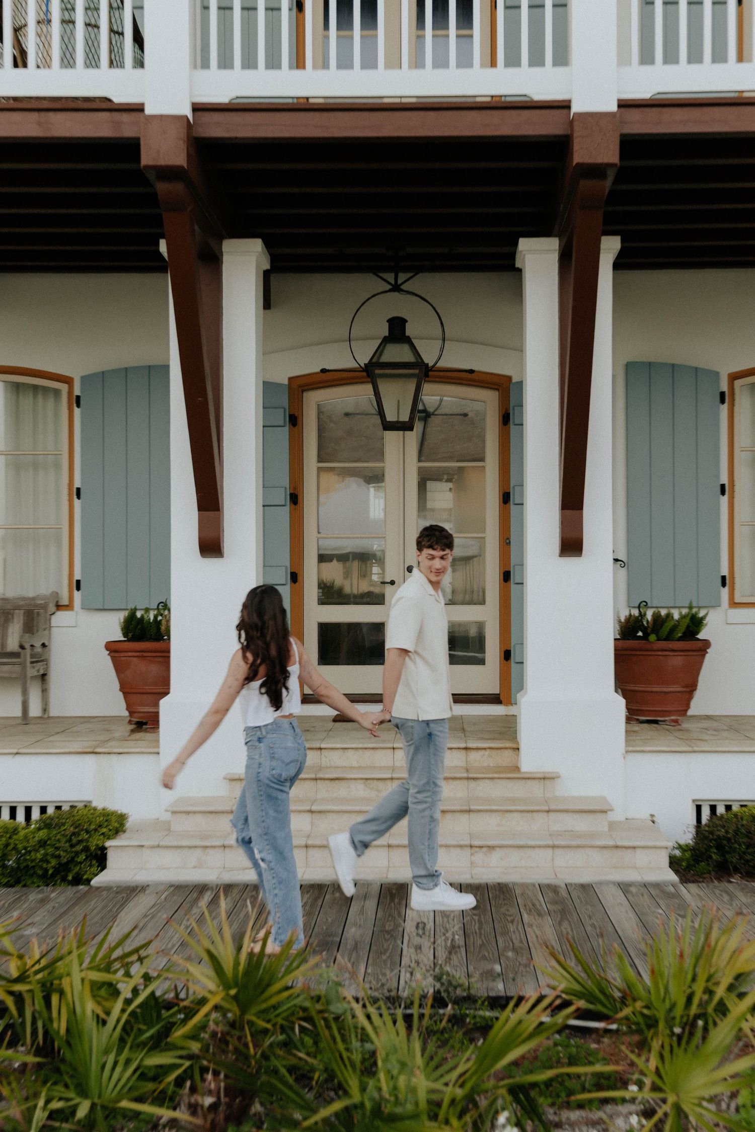 Couple walking together on a white columned porch with potted plants and palm fronds in the foreground.