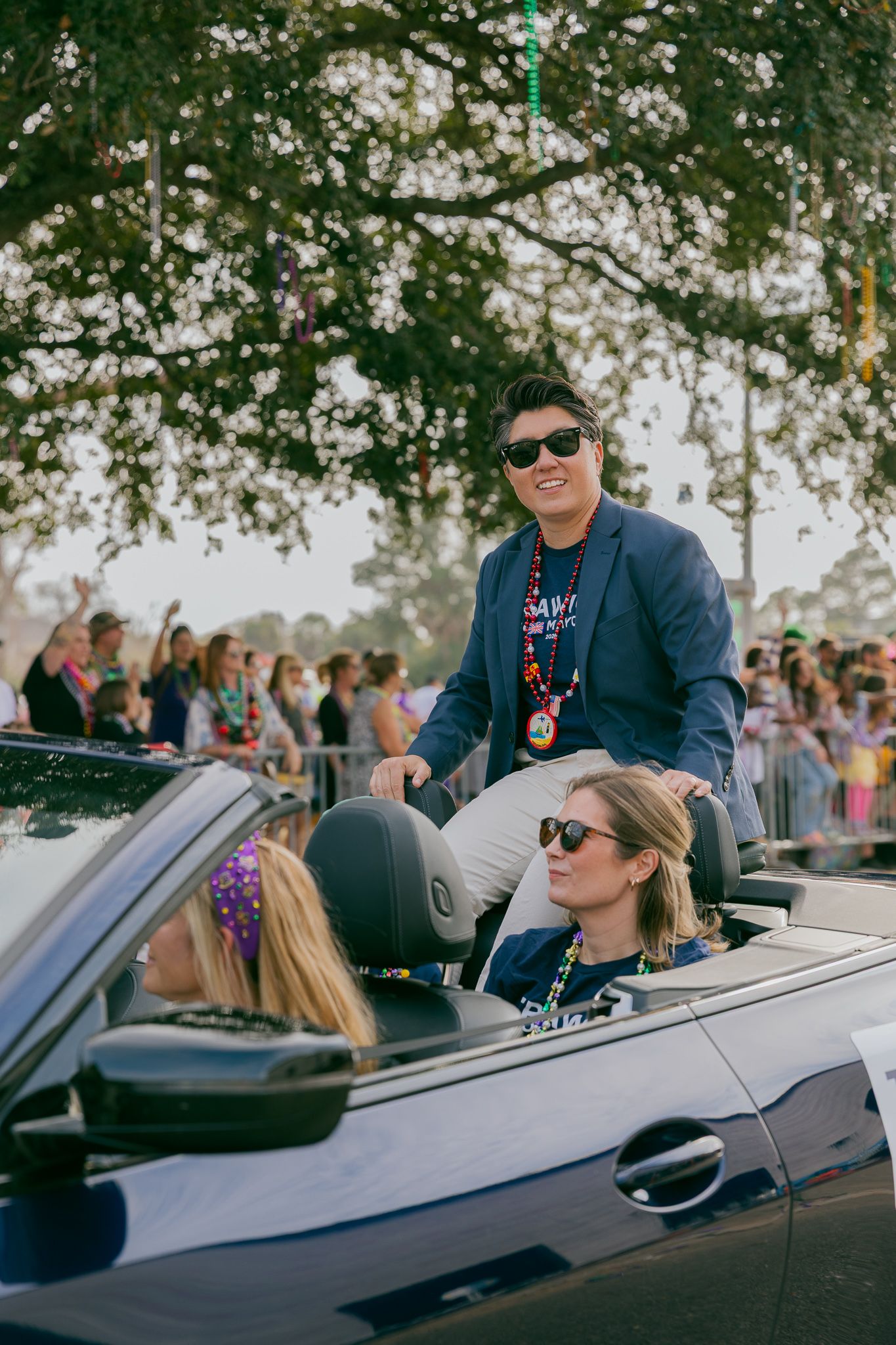 People in sunglasses riding in a convertible car during a sunny outdoor event with spectators visible in background.