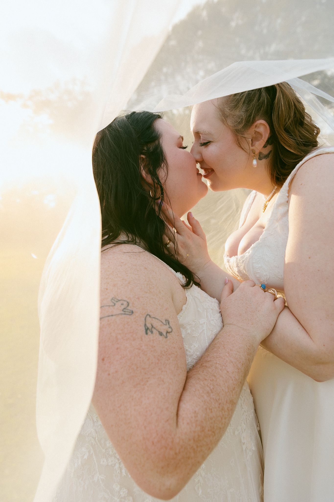 Two brides sharing a romantic kiss under a veil in golden sunlight on their wedding day.