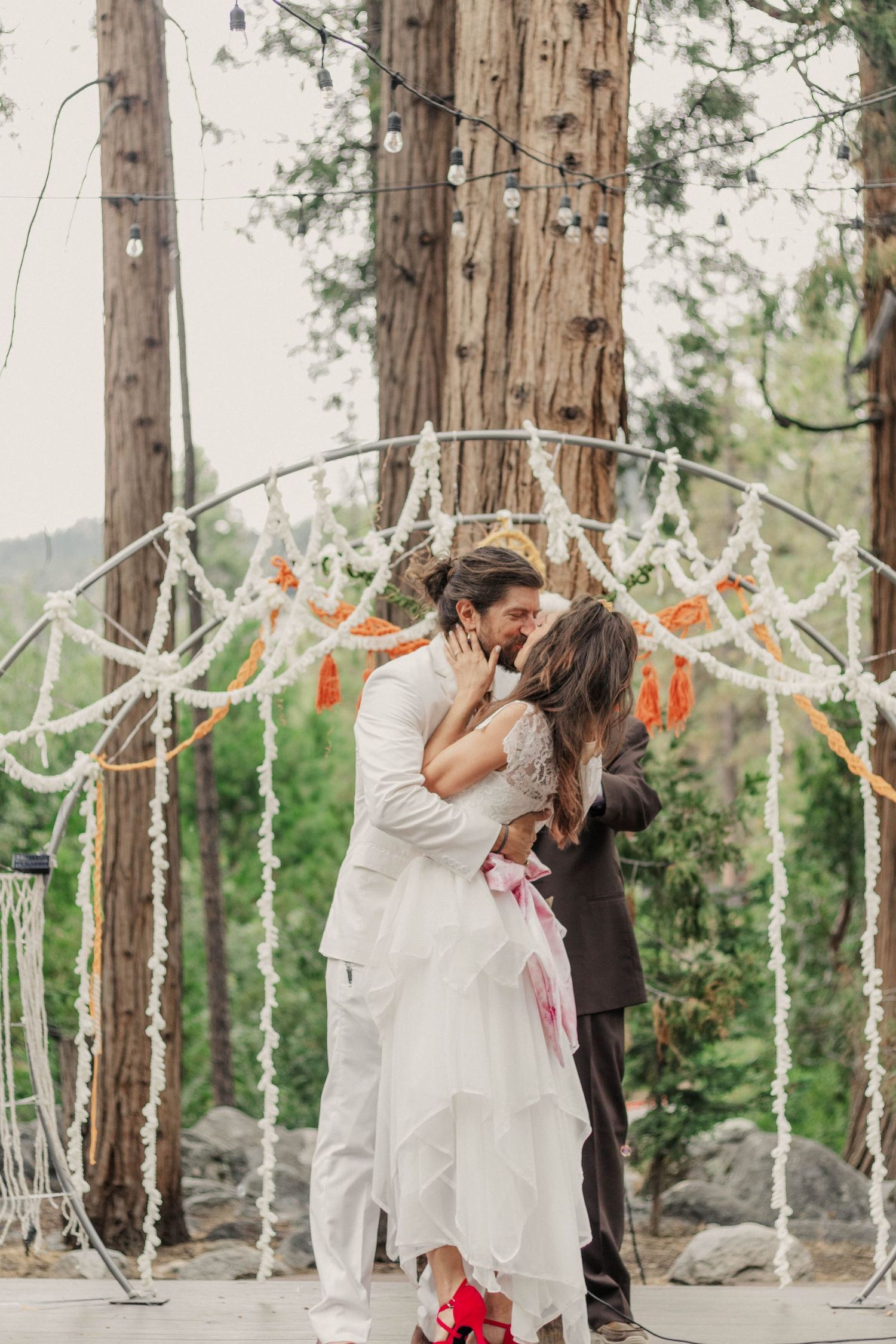 Bride and groom share first kiss at outdoor forest wedding ceremony with boho arch and string lights.