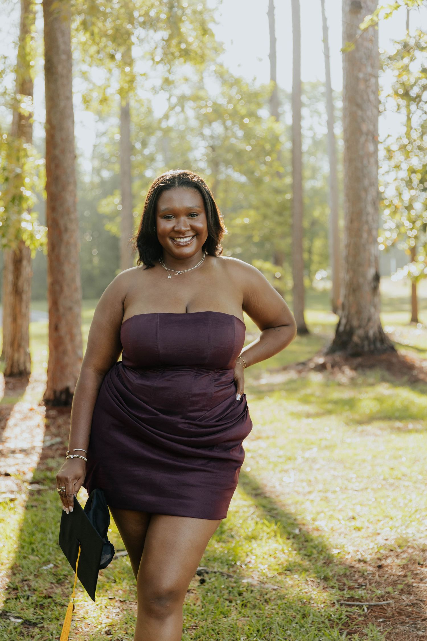 A person in a purple strapless dress poses confidently in a sunlit forest setting.
