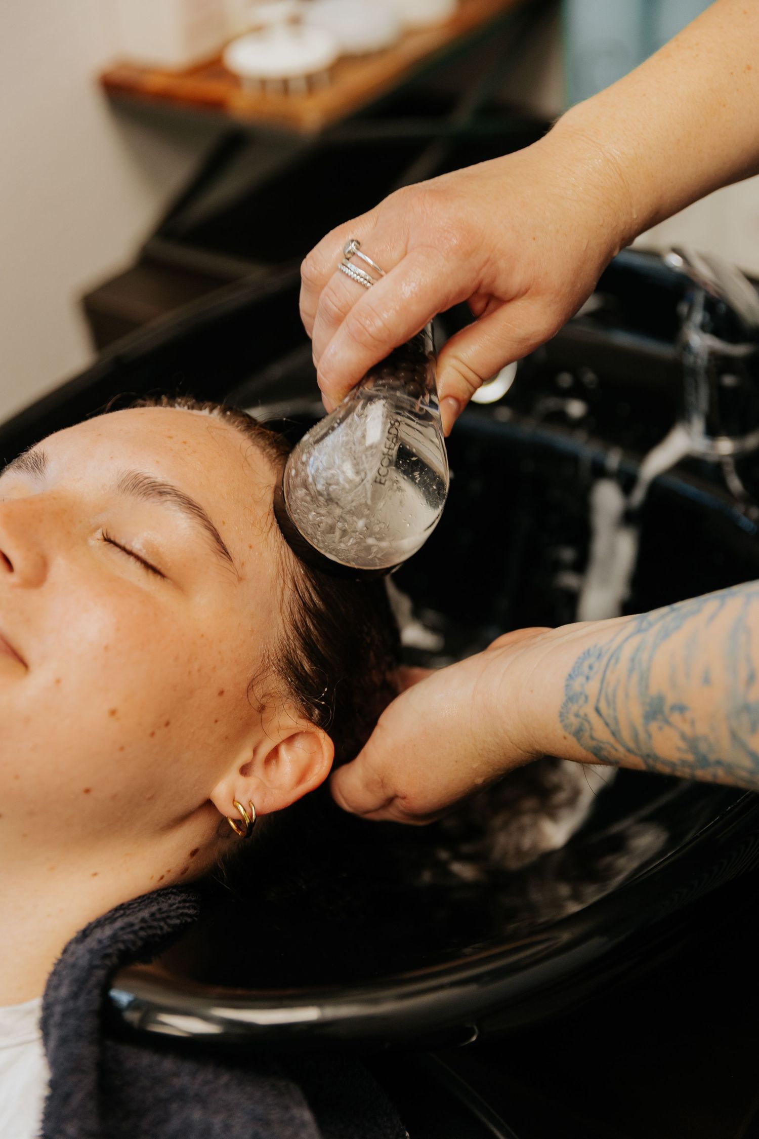 Professional hairstylist washes client's hair at salon sink with black shampoo bowl and tattooed arms visible.