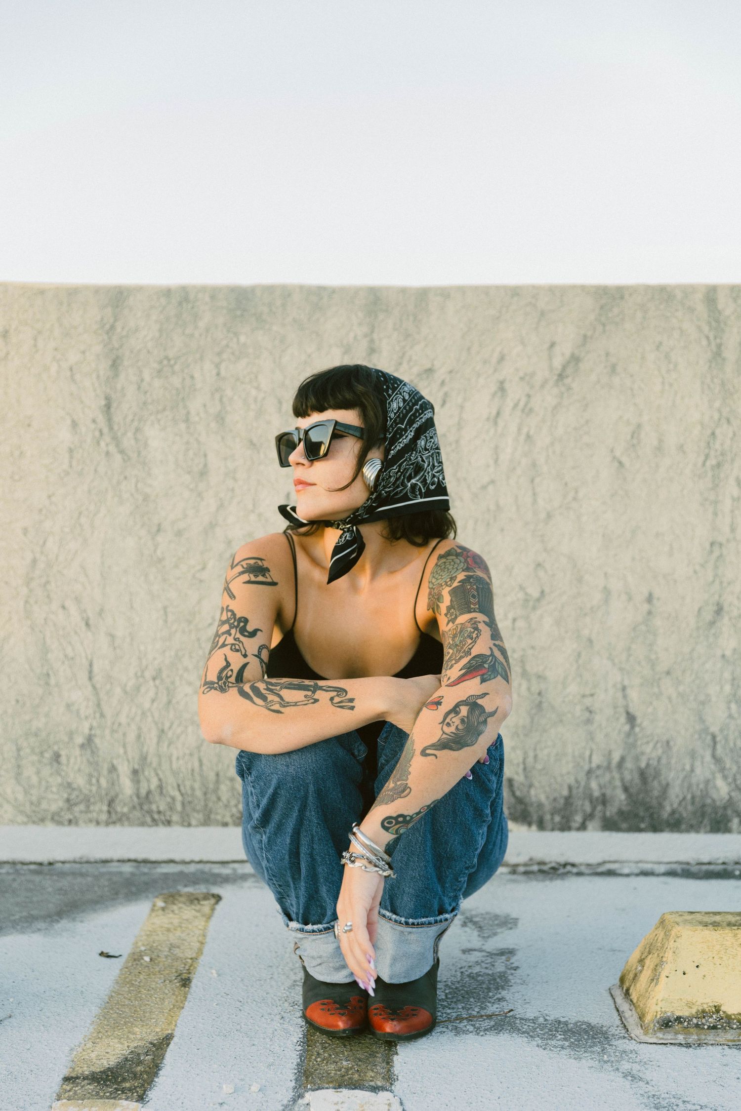 Person with tattoos wearing denim sits crouched against a concrete wall wearing sunglasses.